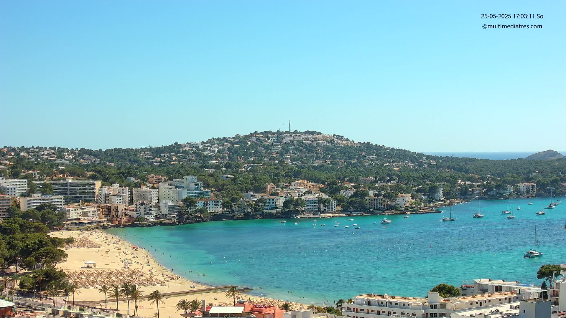 A sunny coastal town with a sandy beach crowded with people and numerous boats in a clear turquoise bay, backed by a hill covered in buildings and trees.