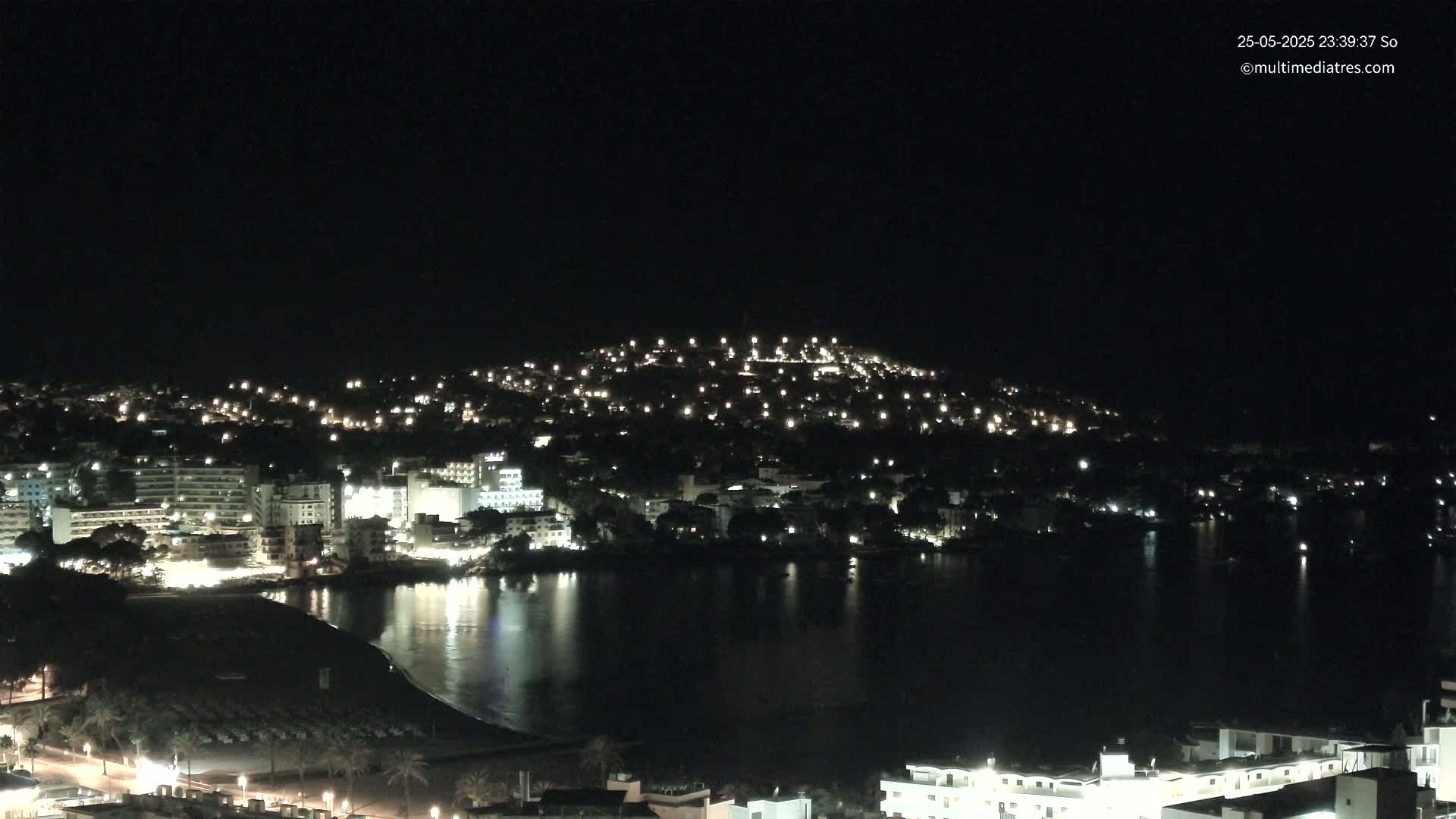 A nighttime view of a coastal town with numerous lights illuminating buildings and a calm body of water reflecting the lights.