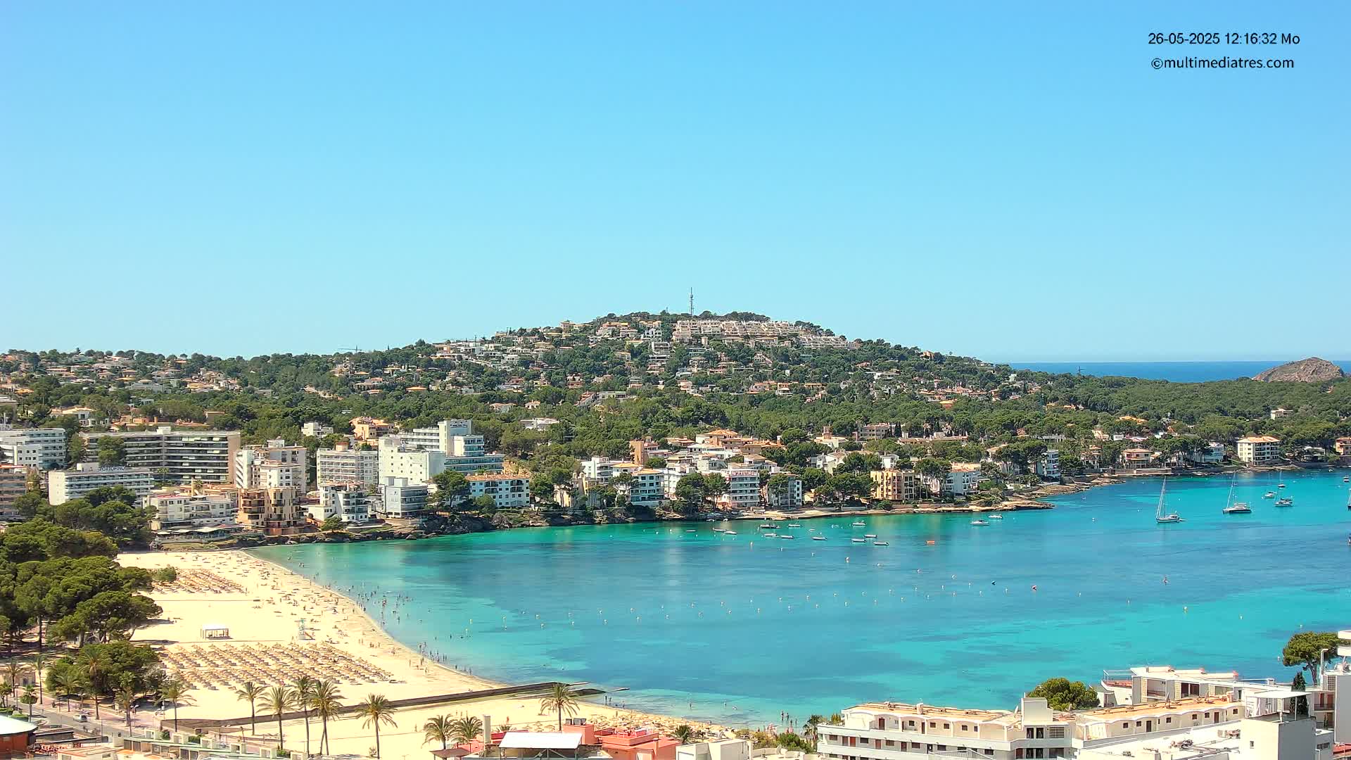 A sunny day reveals a coastal town nestled against a verdant hill, featuring a sandy beach teeming with people and a calm, turquoise bay dotted with sailboats.