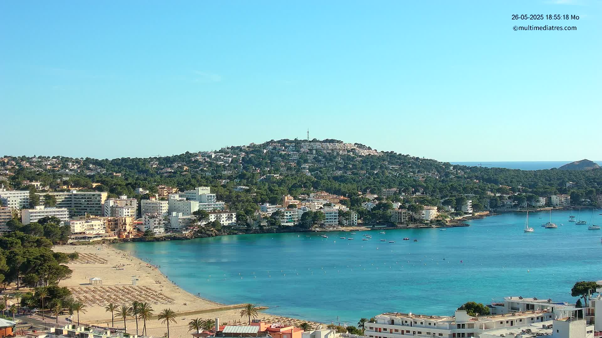 A coastal town nestled against a verdant hill overlooks a calm, turquoise bay under a clear blue sky.