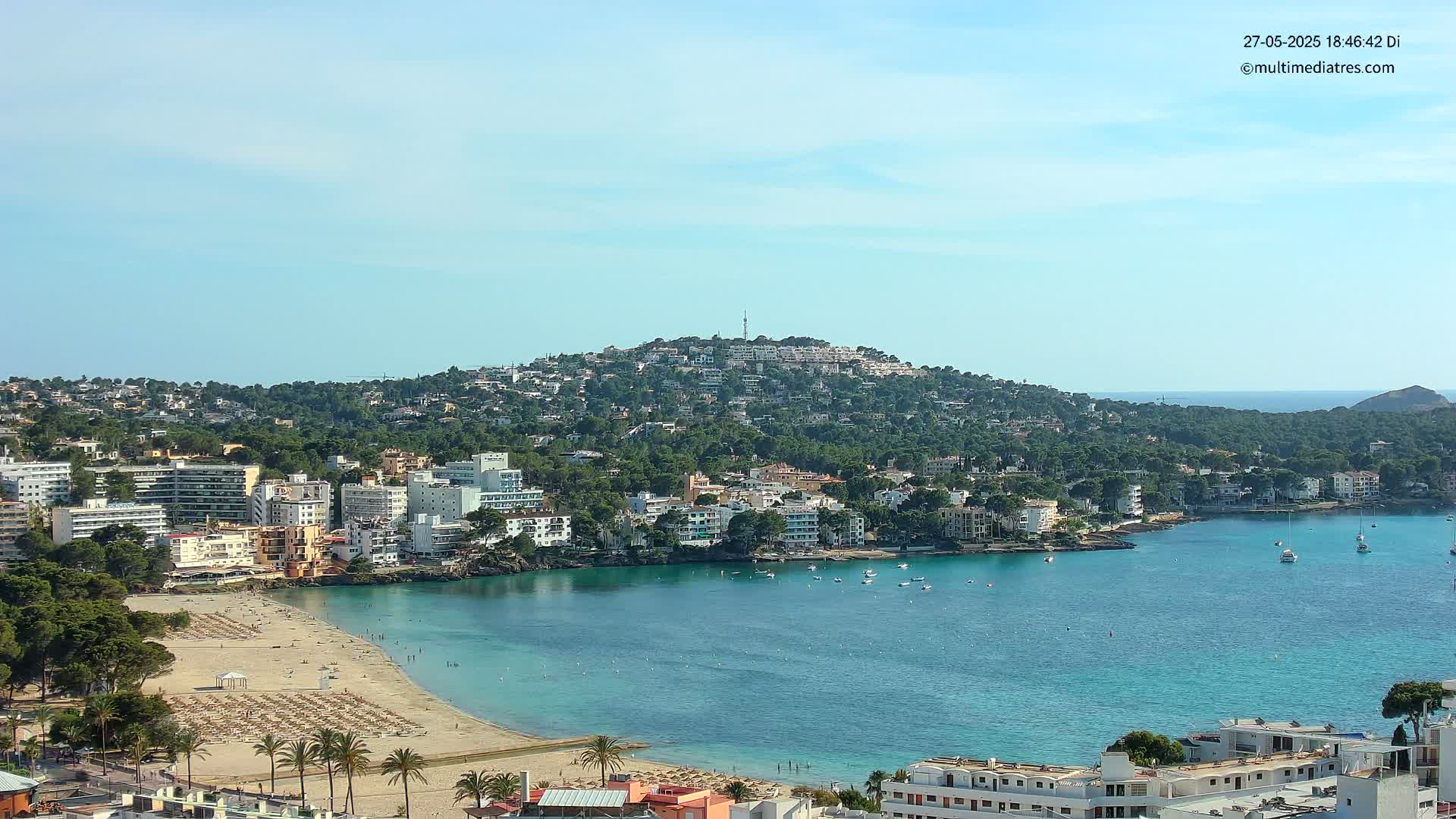 A coastal town nestled against a verdant hill, features a sandy beach with sun loungers and a calm, turquoise bay dotted with boats under a clear sky.