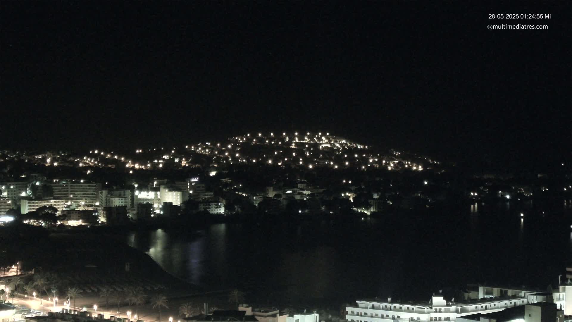 A nighttime view of a coastal town illuminated by many lights, with a dark body of water in the foreground under a clear sky.