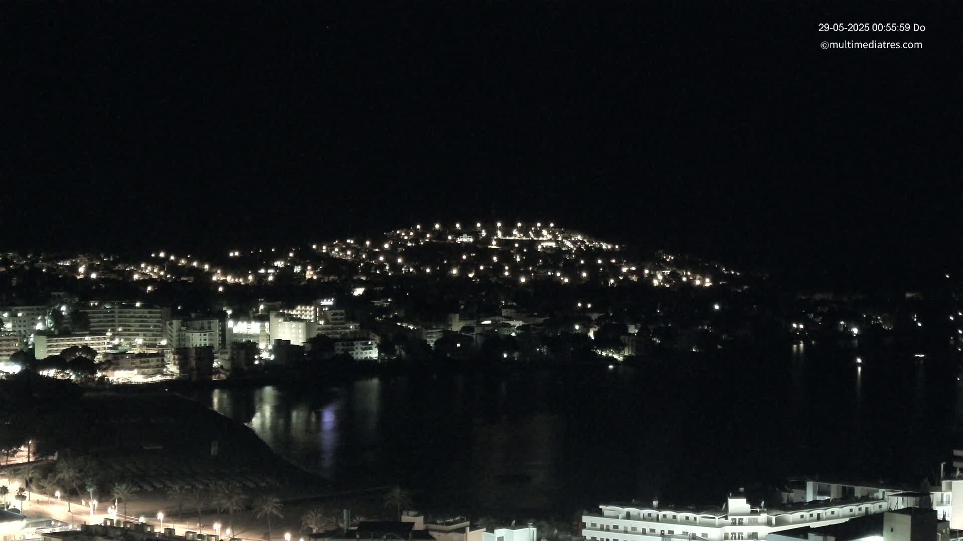 A nighttime view of a coastal town illuminated by many lights, with calm dark water reflecting the lights in the foreground.