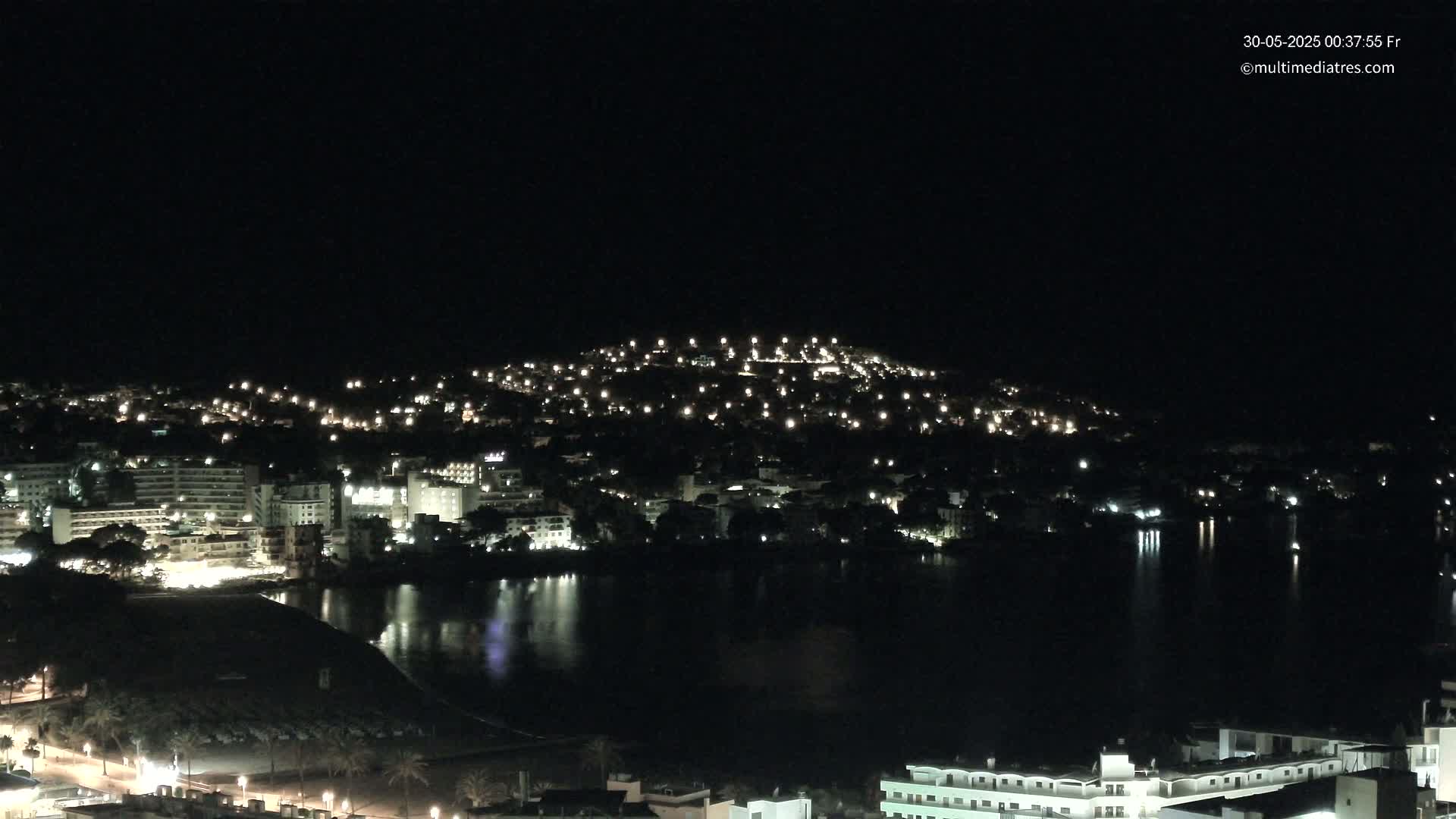A nighttime aerial view shows a coastal town with many lights illuminating buildings and streets along a dark body of water.