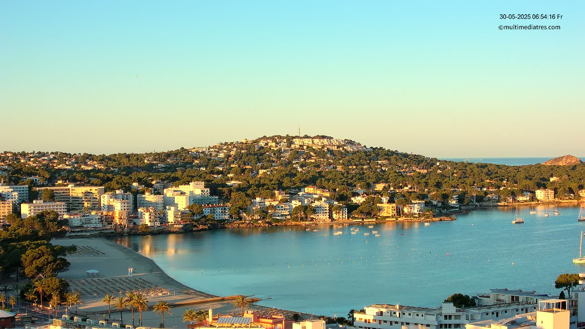A coastal town nestled on a hillside, with a calm bay featuring numerous boats and a sandy beach under a clear, light-blue sky.