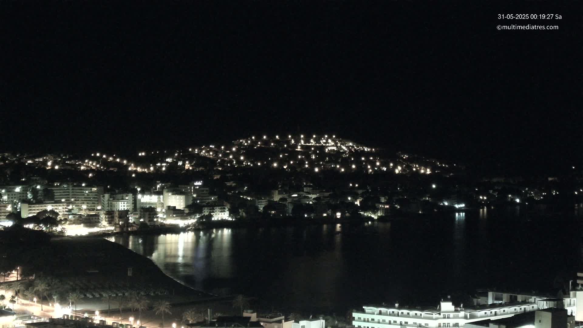 A nighttime view of a coastal town illuminated by lights, with calm dark water reflecting the lights in front of it.