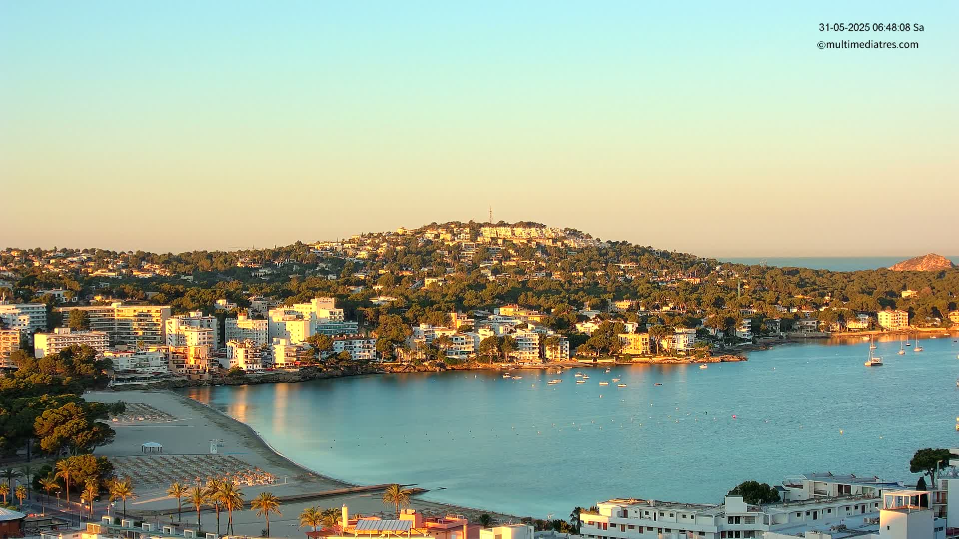 A coastal town nestled against a hill, with a calm bay and sandy beach, under a clear sunrise sky.