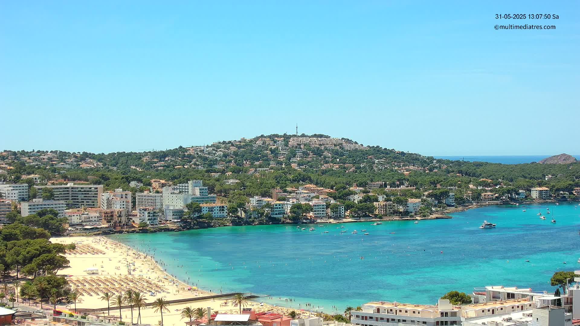 A sunny, clear day reveals a coastal town nestled against a verdant hill overlooking a calm, turquoise bay with a sandy beach filled with sunbathers and boats in the water.