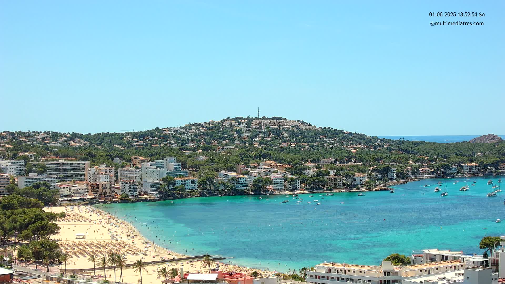 A sunny day shows a crowded beach, turquoise bay filled with boats, and a hillside town under a clear blue sky.