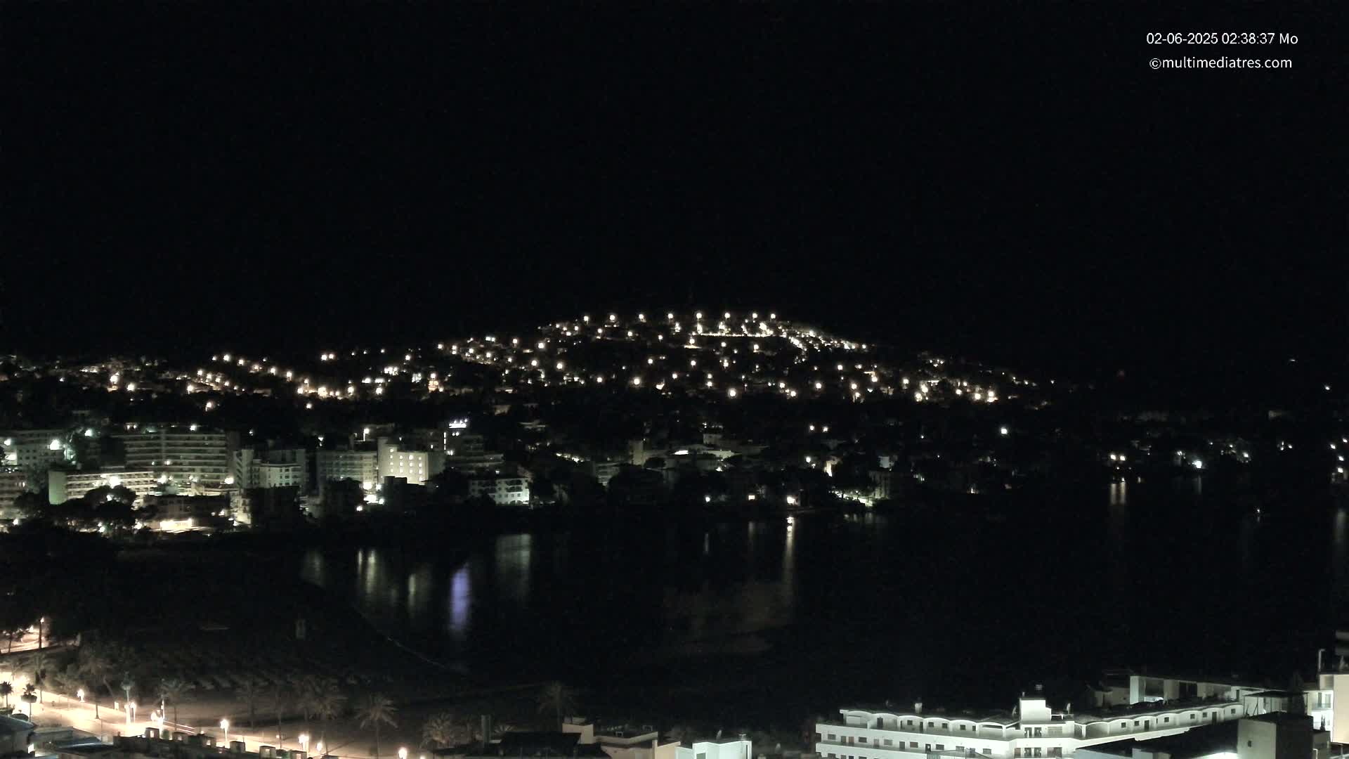 A nighttime view of a coastal town with buildings illuminated, bordering a dark body of water under a clear, dark sky.