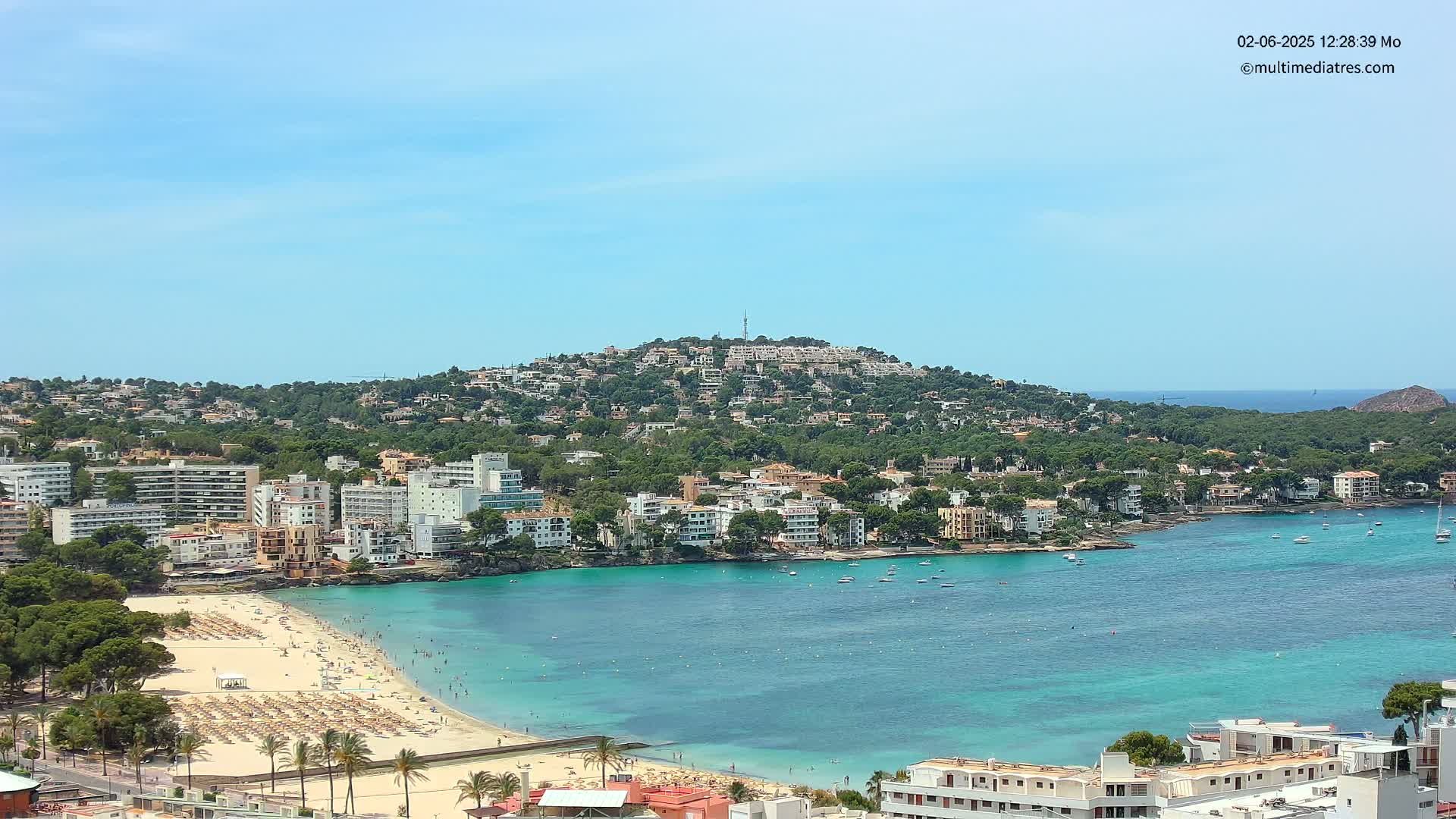 A sunny coastal town with a sandy beach, turquoise water, and numerous buildings nestled on a hillside overlooking a calm bay.