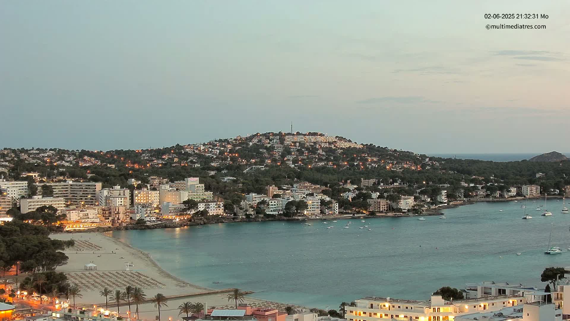 A coastal town at twilight, nestled around a calm bay with a hilltop illuminated at night.