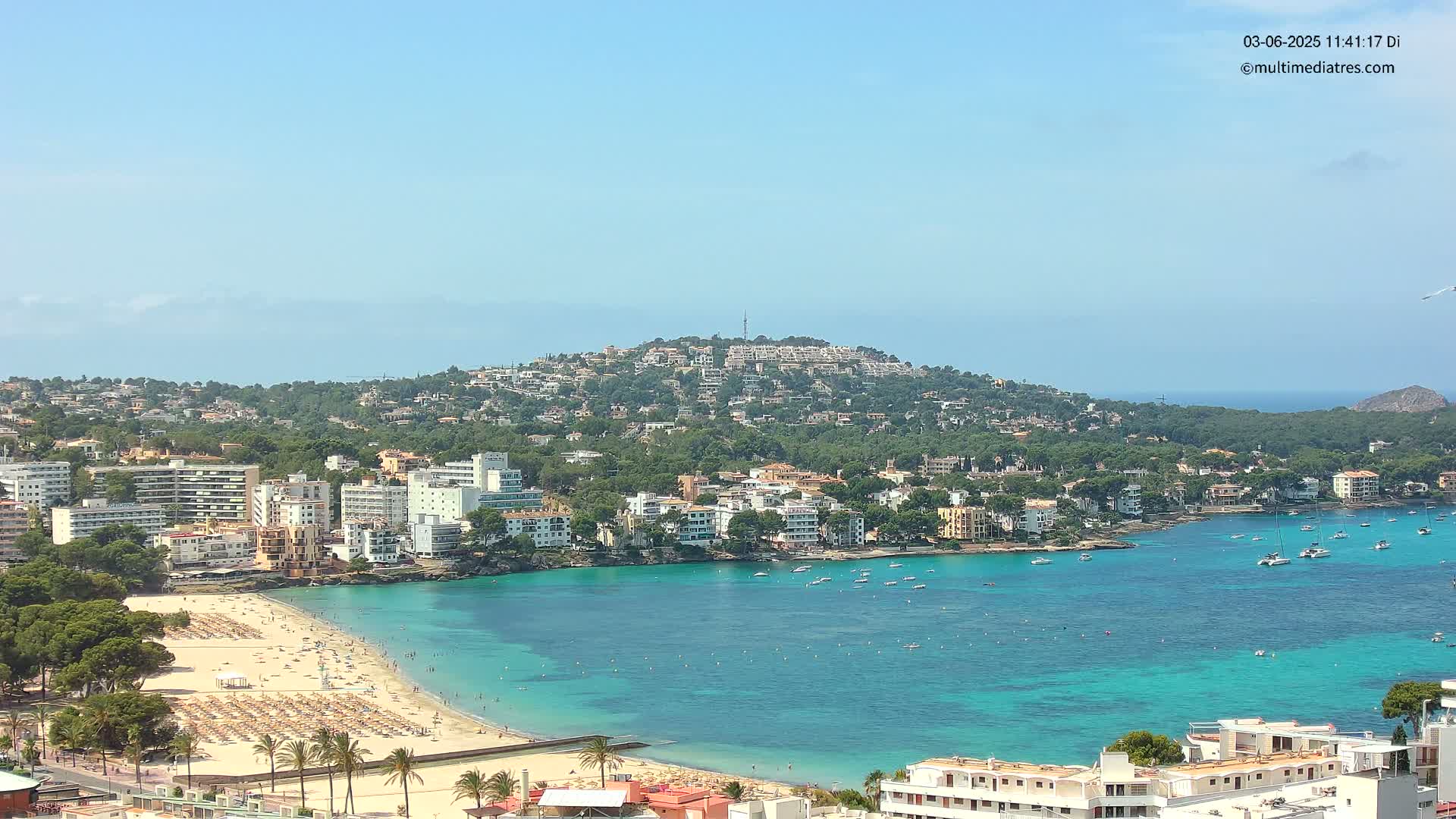 A sunny day reveals a coastal town nestled against a hill overlooking a turquoise bay with a sandy beach and numerous boats.