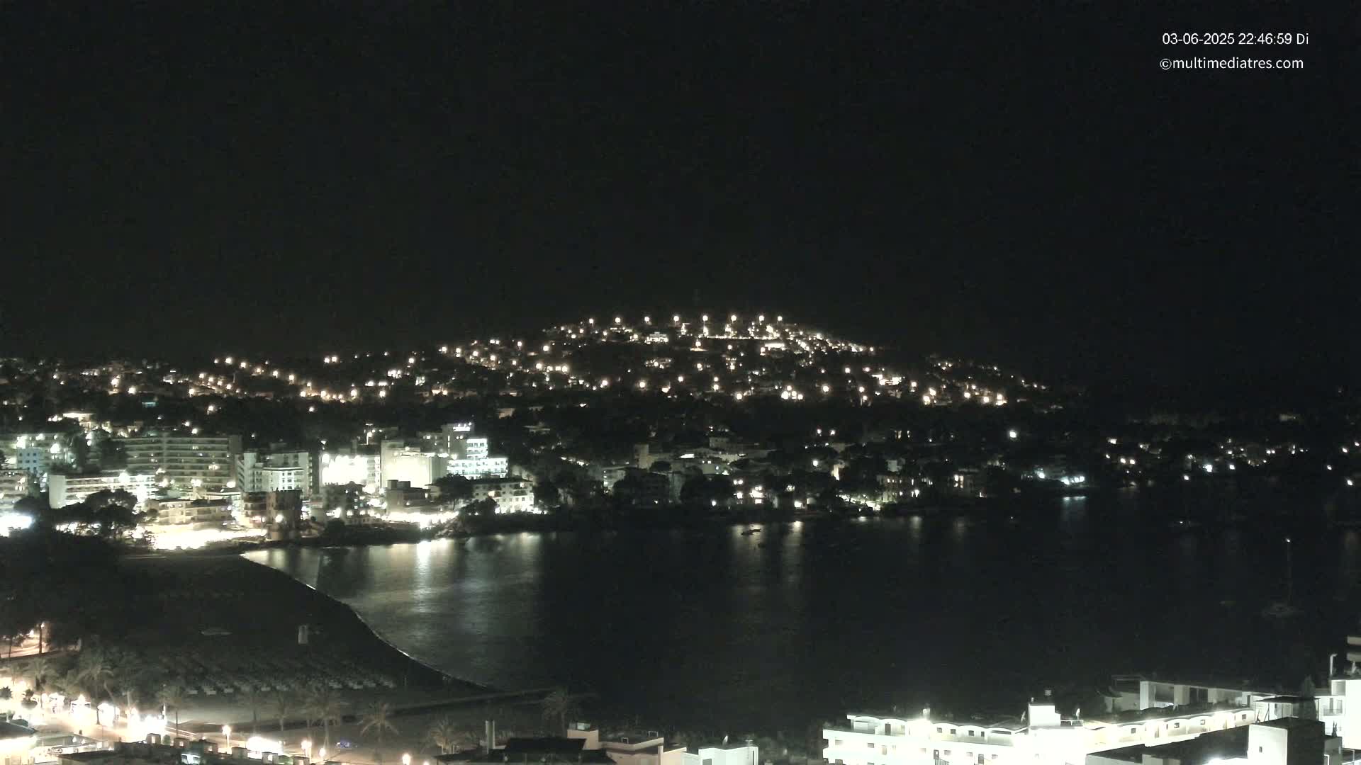 A nighttime, aerial view shows a coastal town illuminated by lights, with a dark body of water separating the town from a hill also dotted with lights.