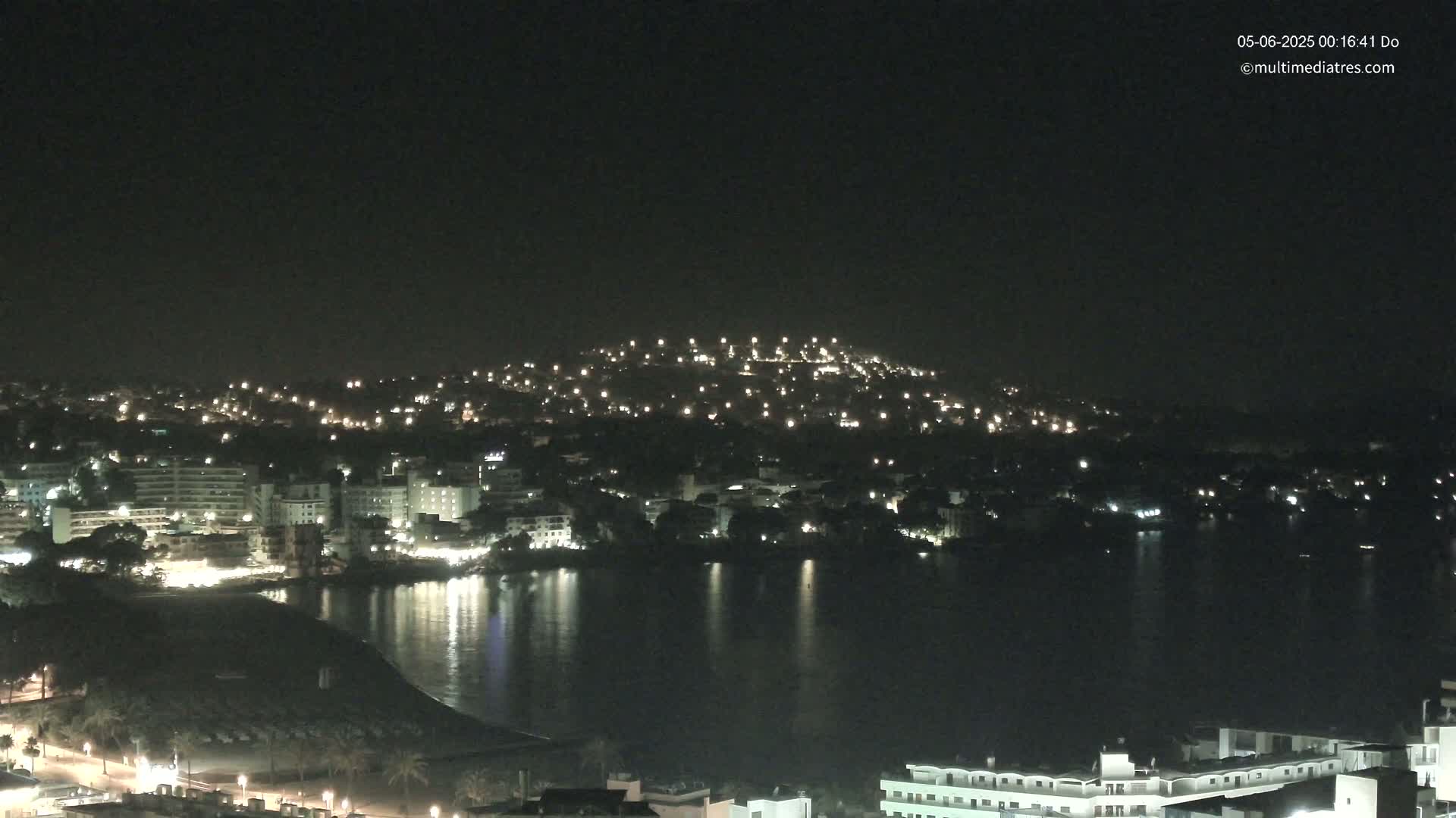 A nighttime view of a coastal town with many lights illuminating buildings and a calm body of water reflecting the lights.