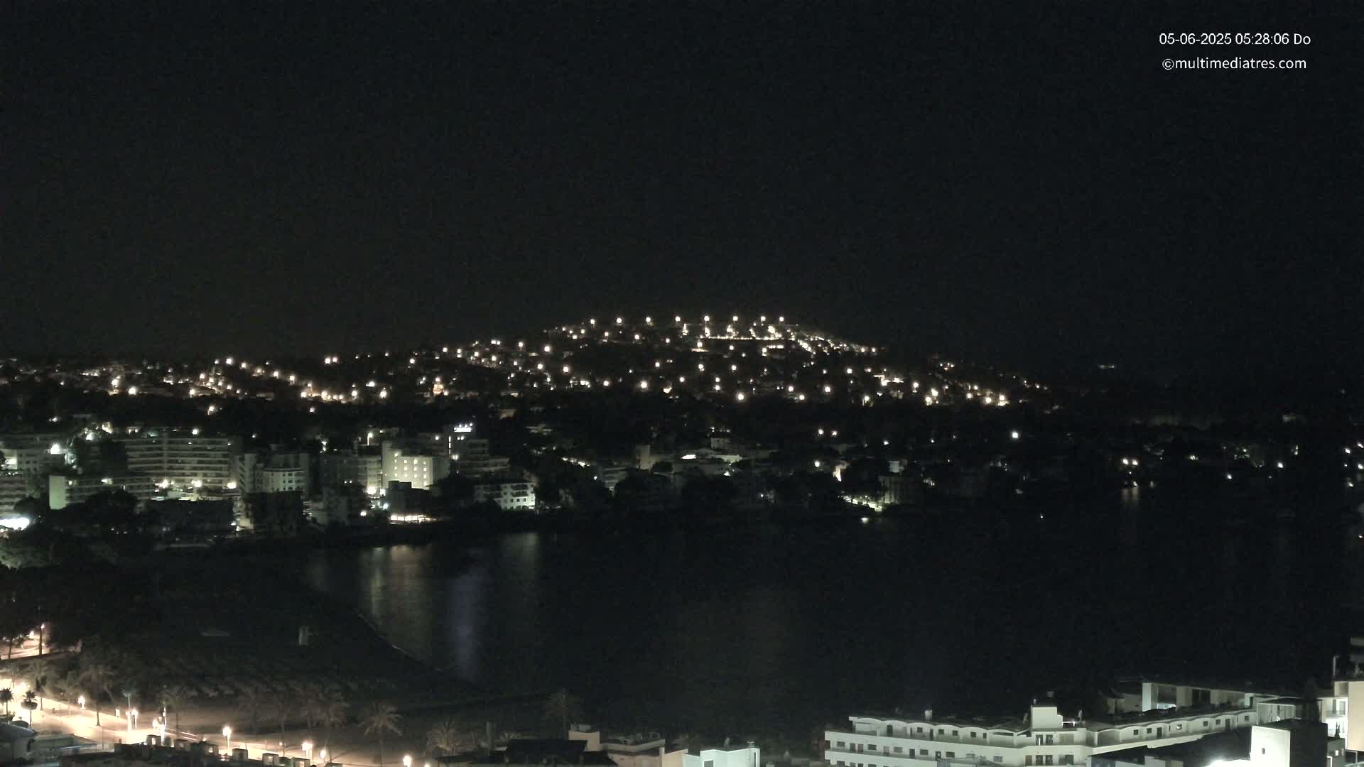 A nighttime view of a coastal town illuminated by many small lights, situated on a dark hill overlooking a body of dark water.