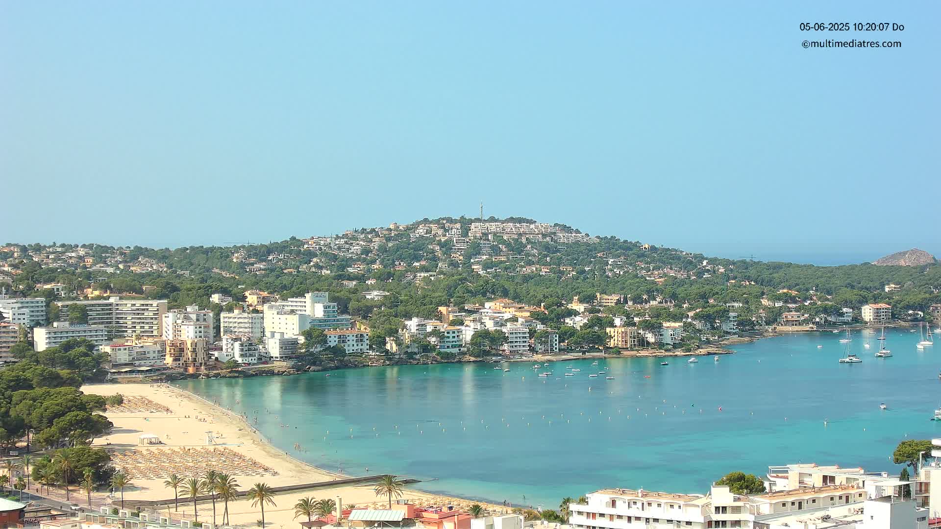 A coastal town with a sandy beach and turquoise water is nestled beneath a hill covered in buildings and vegetation under a clear, bright sky.