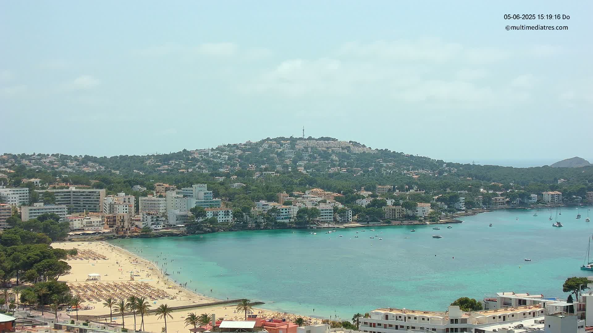 A sunny day shows a coastal town nestled beside a calm, turquoise bay, with a hill densely covered in buildings and trees in the background.