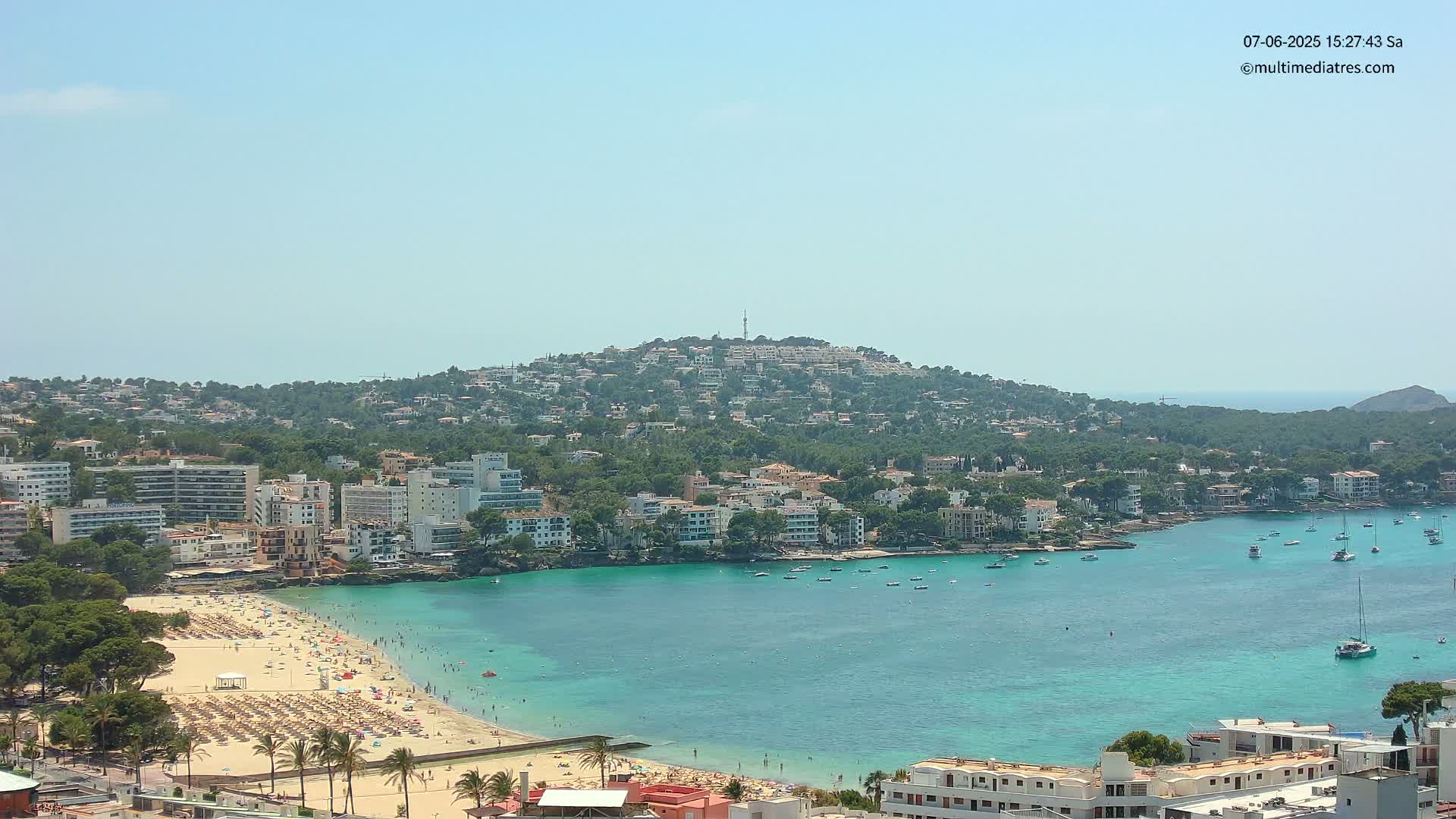 A sunny coastal town with a sandy beach crowded with people and numerous buildings extending up a hillside, all nestled in a turquoise bay dotted with boats.