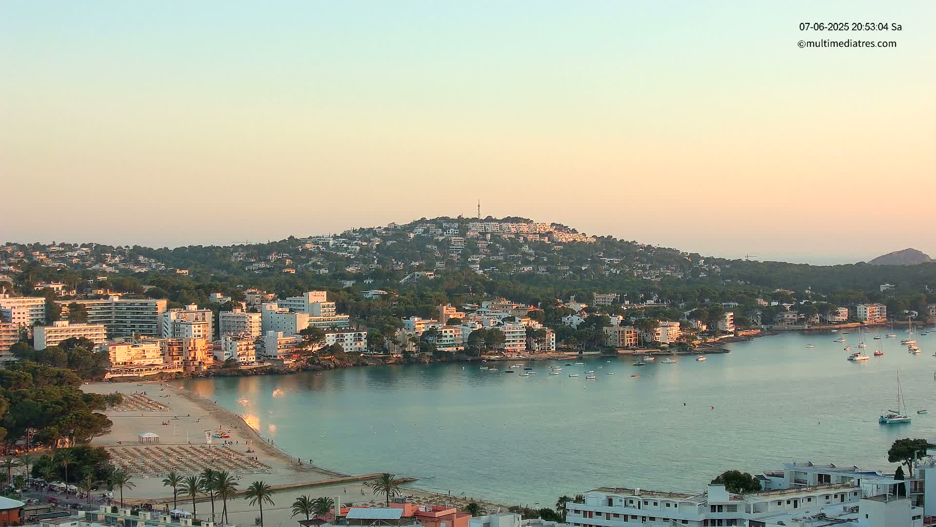 A coastal town with a sandy beach and calm, turquoise waters is nestled at the base of a hill covered in buildings and lush greenery under a pale orange sunset sky.
