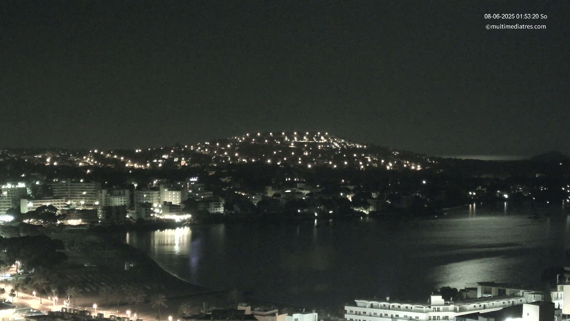 A nighttime view of a coastal town with a calm body of water reflecting the lights from buildings on a hill and along the shoreline.