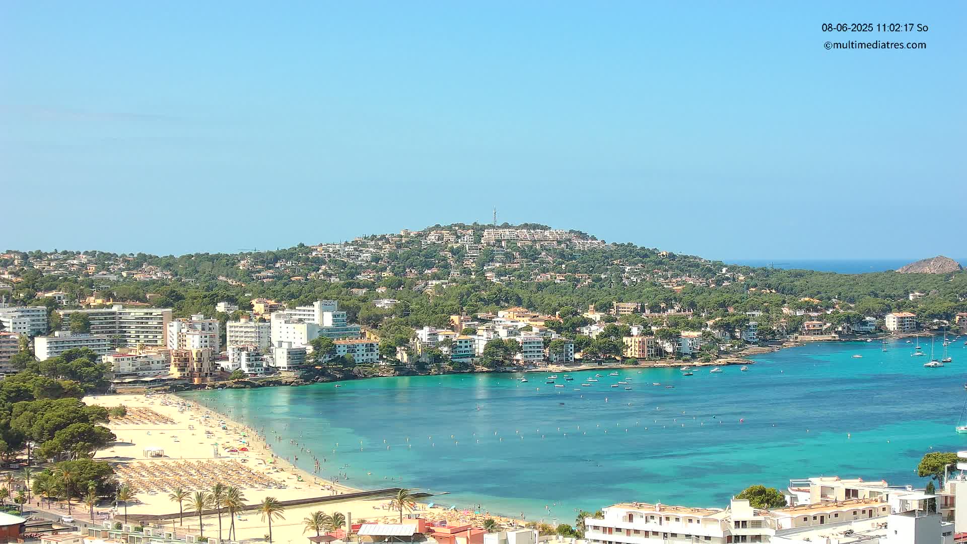 A clear, sunny day overlooks a coastal town with a sandy beach, turquoise water filled with boats, and buildings covering a hillside.