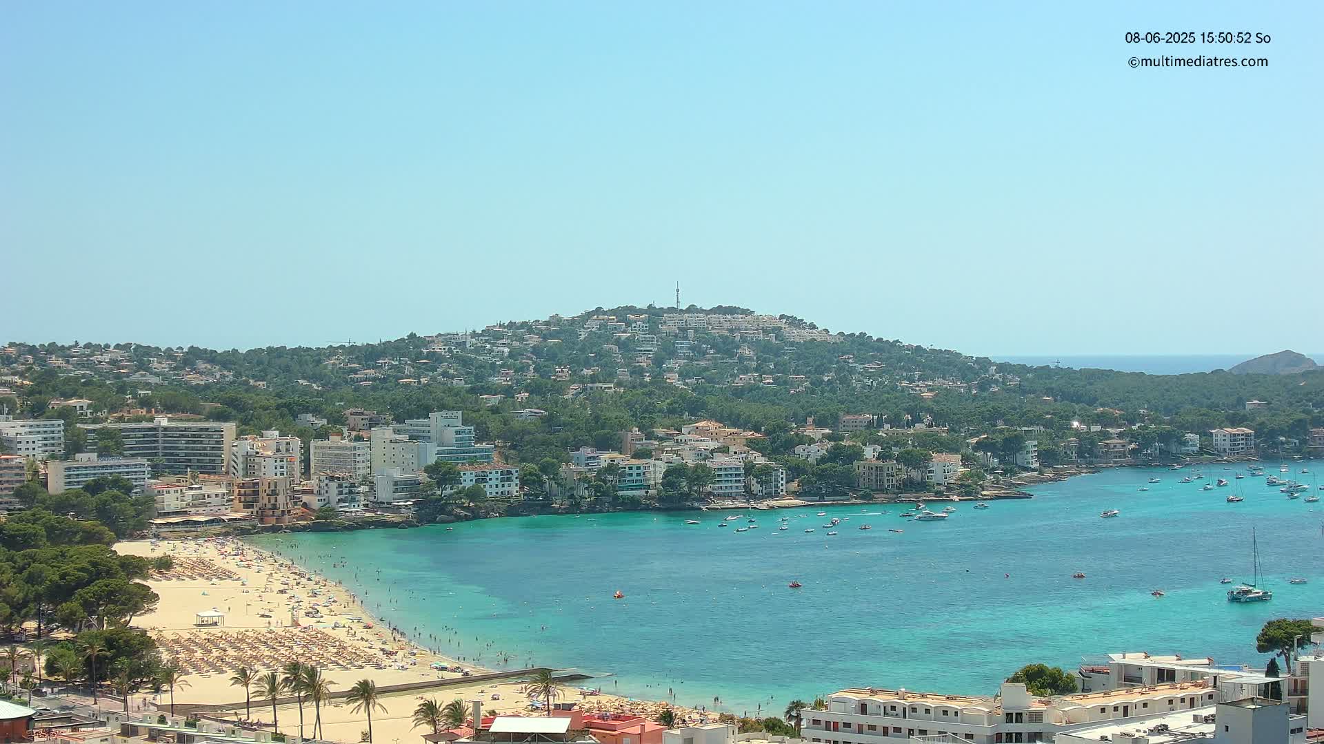 A sunny day reveals a coastal town nestled against a verdant hill, featuring a sandy beach crowded with sunbathers and a calm turquoise bay dotted with boats.