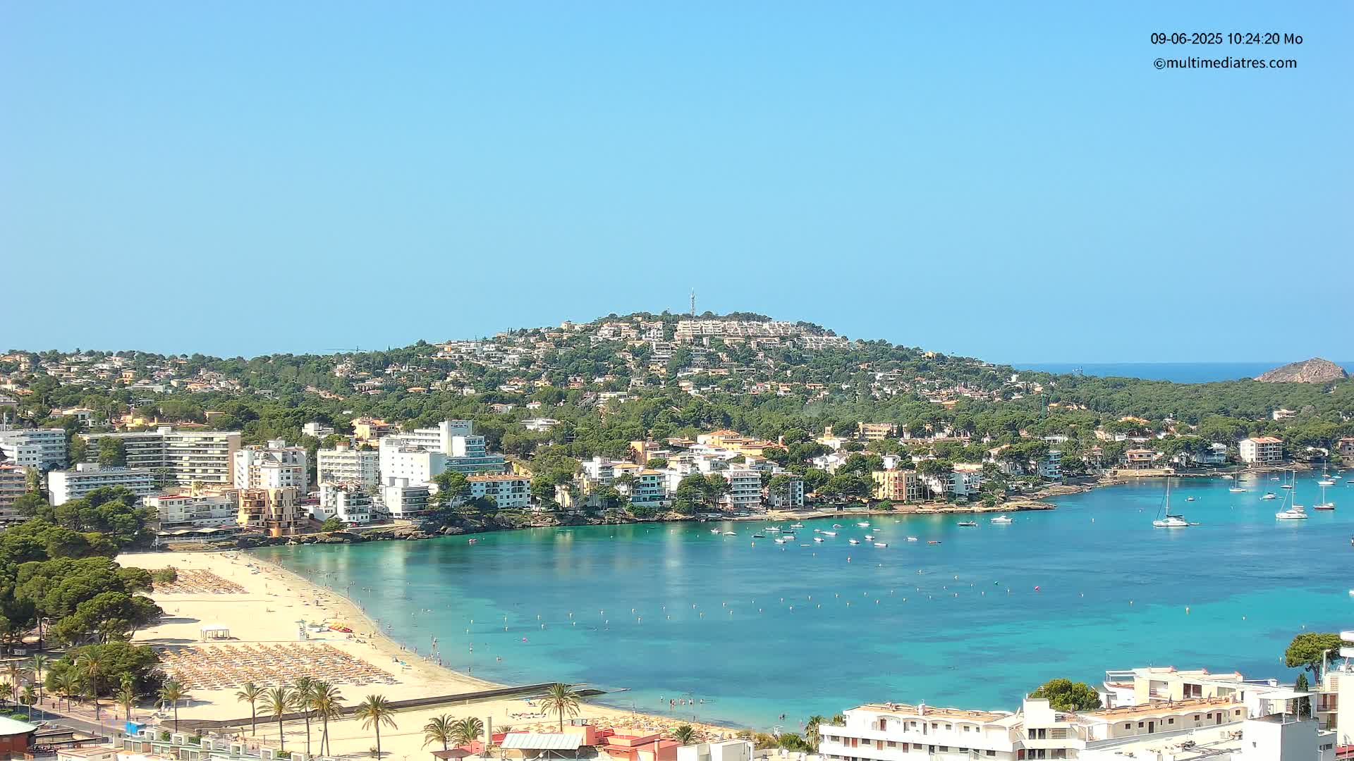 A sunny coastal town with a sandy beach and turquoise bay, nestled against a hillside covered in buildings and trees.