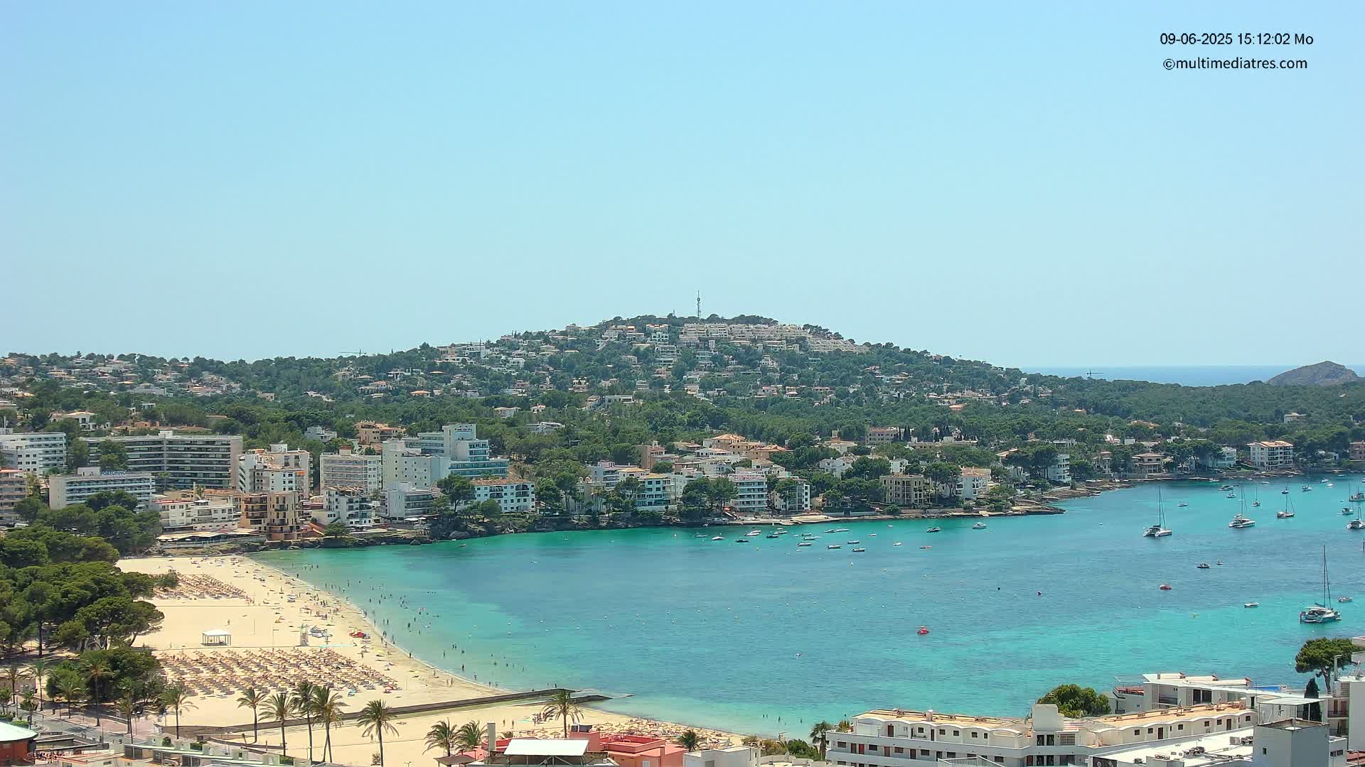 A sunny coastal town with a sandy beach, turquoise water, and numerous buildings sits nestled against a lush green hill.