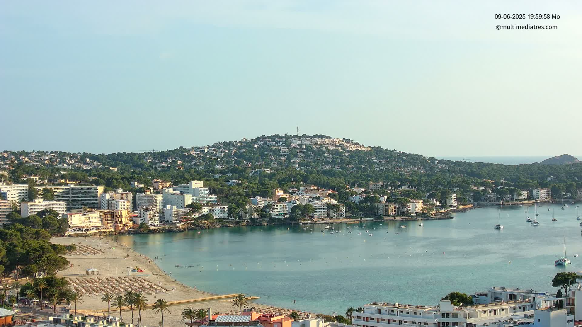A coastal town with buildings and a sandy beach is nestled below a hill covered in trees and buildings, all under a clear sky.