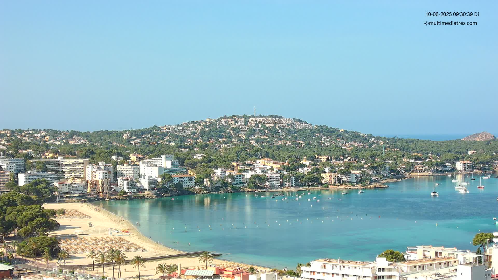 A coastal town nestled on a hillside, with a sandy beach and calm, turquoise water filled with boats, under a clear blue sky.