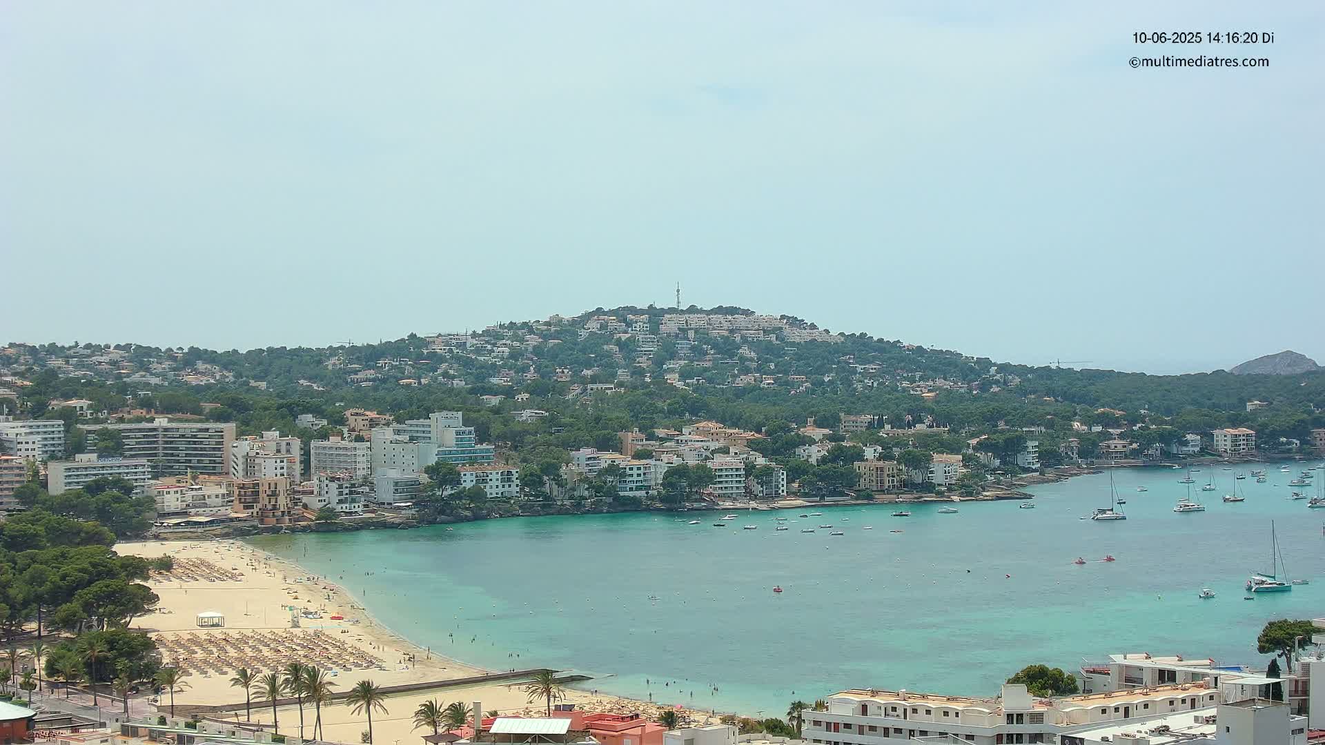 A sunny coastal town with a sandy beach, turquoise water filled with boats, and buildings covering a hillside.