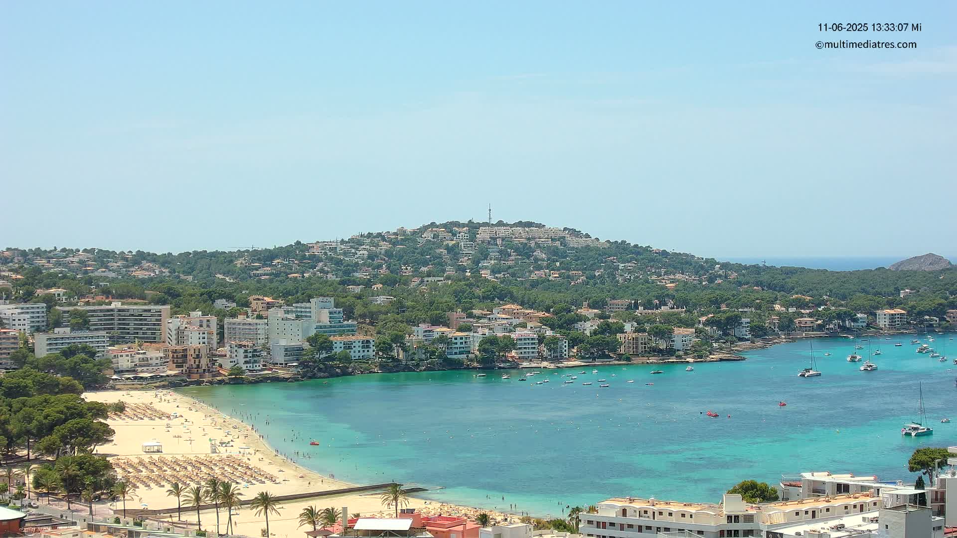 A sunny day reveals a coastal town nestled around a turquoise bay, with a hill covered in houses and trees rising in the background.