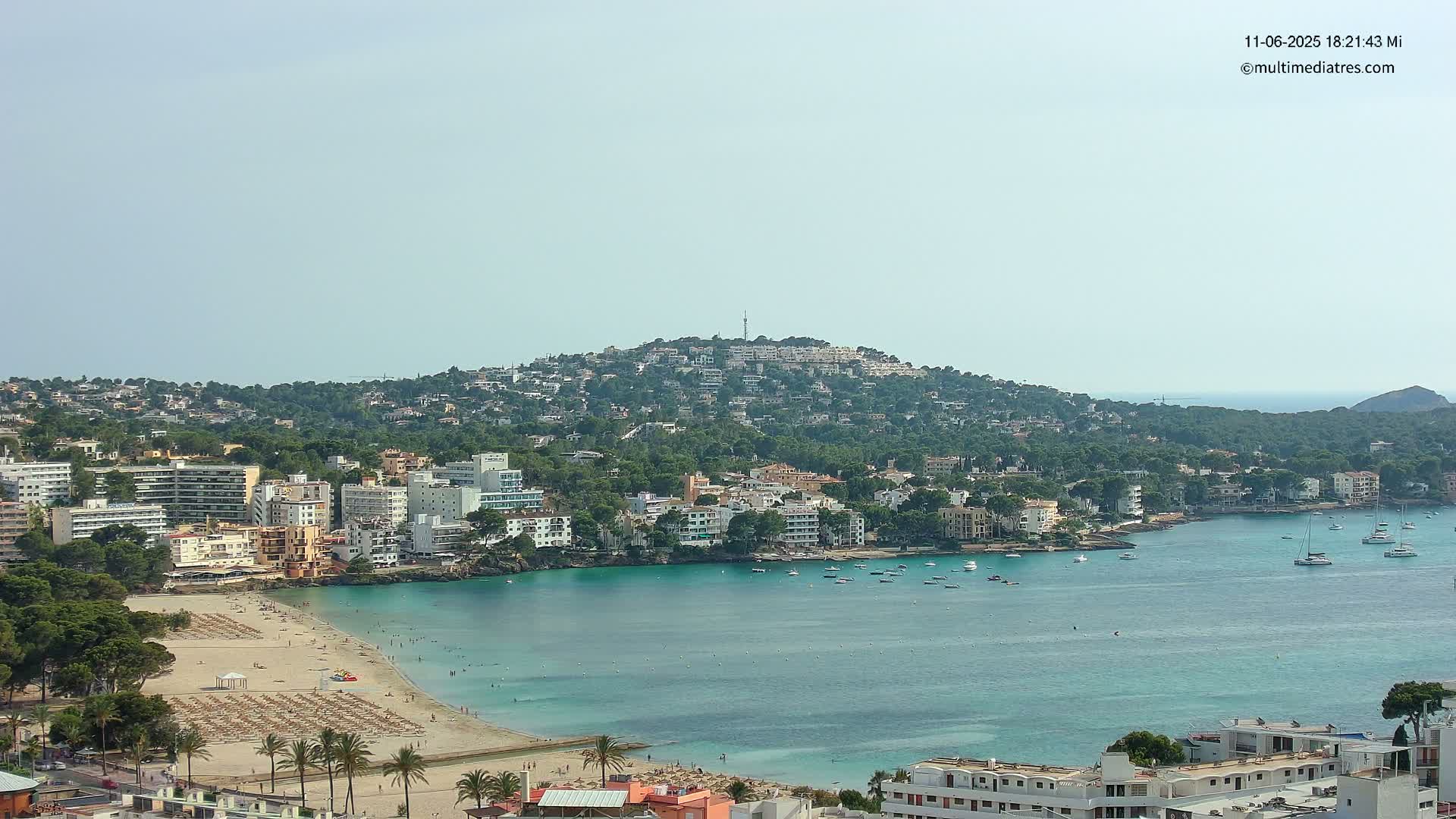 A coastal town with a sandy beach and turquoise water sits below a hill covered in buildings and greenery, under a clear sky.