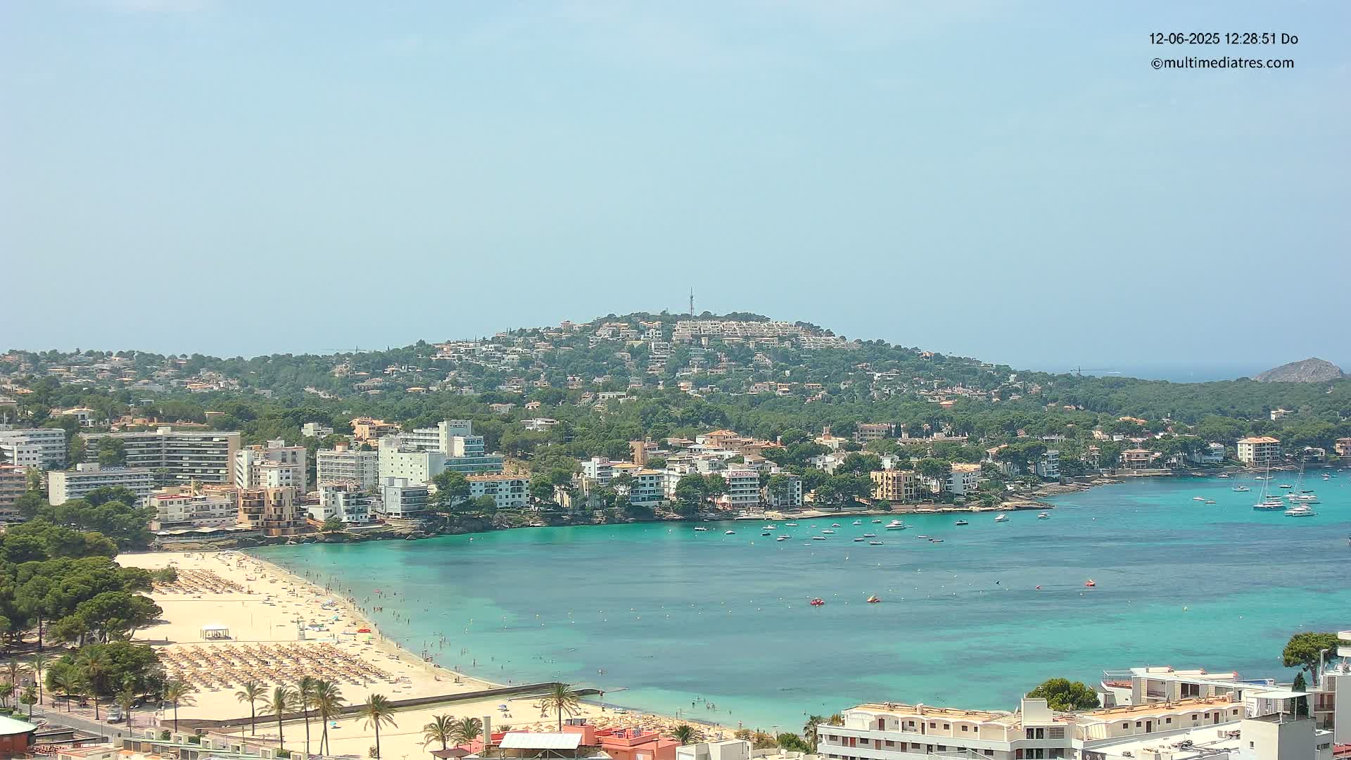 A sunny day reveals a coastal town nestled against a verdant hill, its turquoise bay dotted with boats and a sandy beach crowded with people.