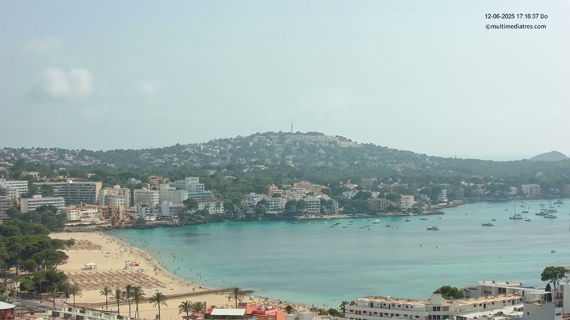 A hazy, sunny day overlooks a coastal town nestled beside a calm, turquoise bay with a sandy beach dotted with umbrellas and people, and a hill covered in buildings and trees in the background.