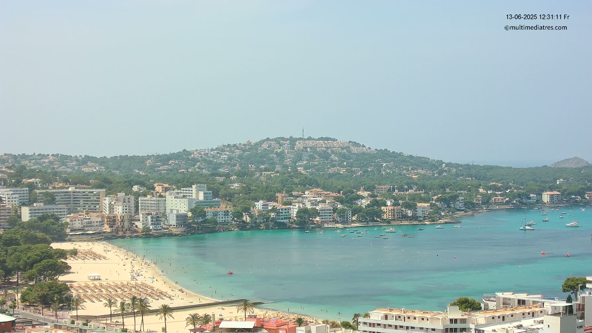 A hazy, sunny day overlooks a coastal town with a sandy beach, turquoise water filled with boats, and buildings that cover a hill in the background.