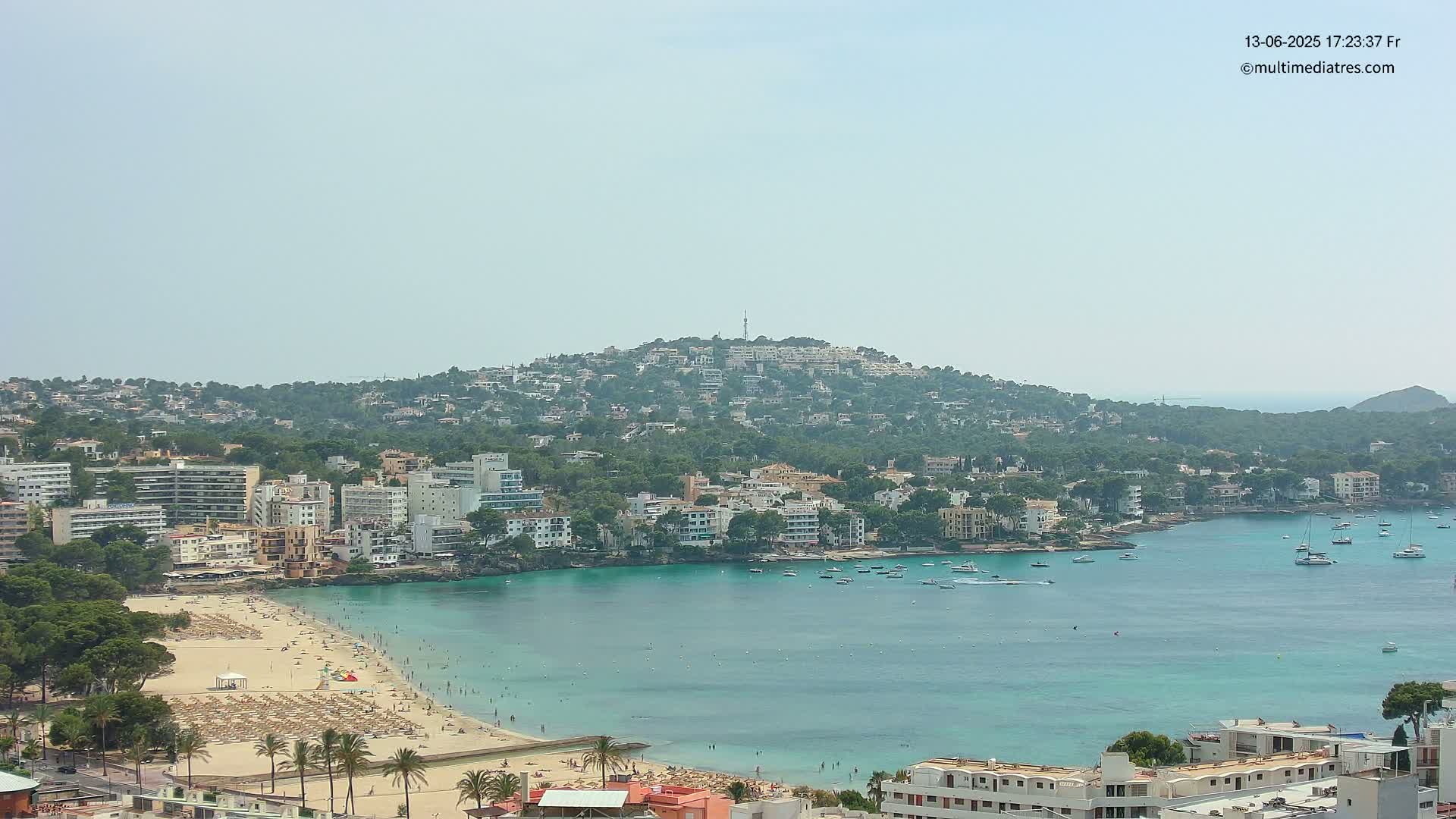 A hazy, sunny day reveals a coastal town nestled along a turquoise bay, with a hill dotted with buildings rising in the background.