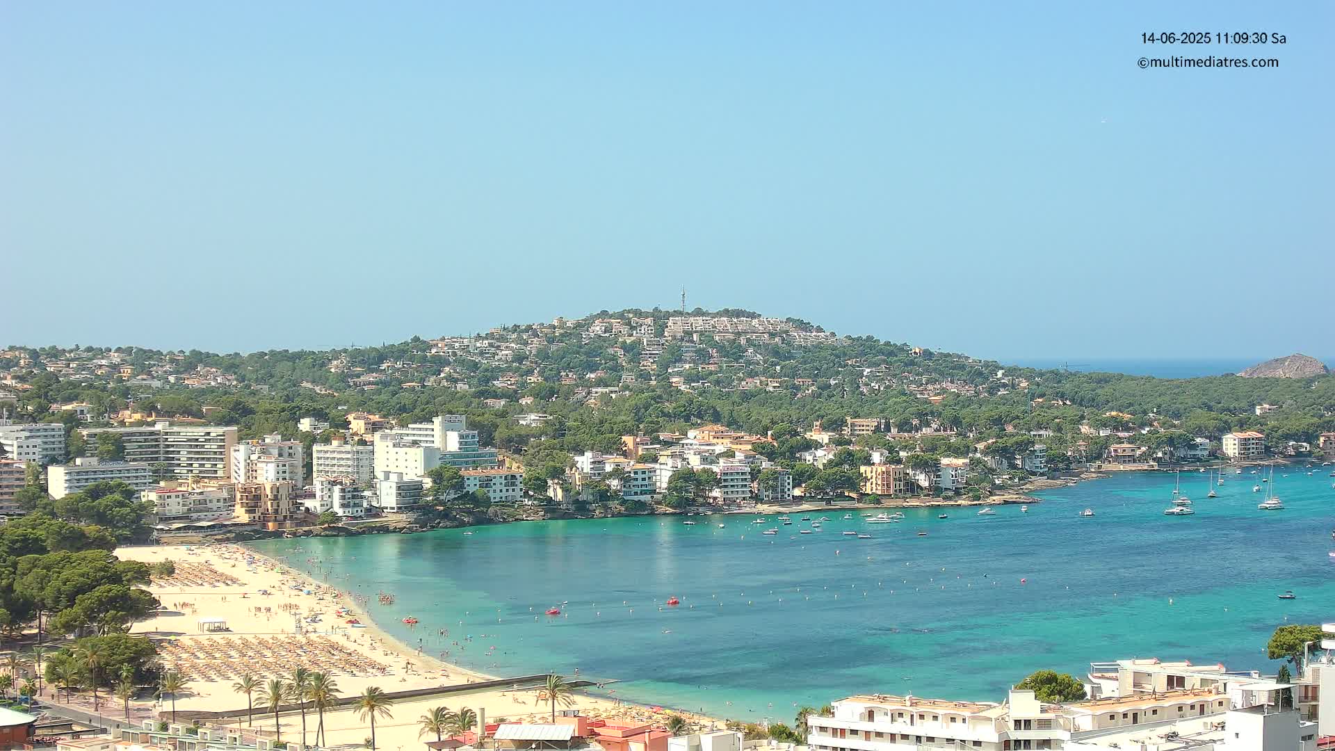 A sunny coastal town with a crowded beach and turquoise water, nestled against a hill covered in buildings and trees.