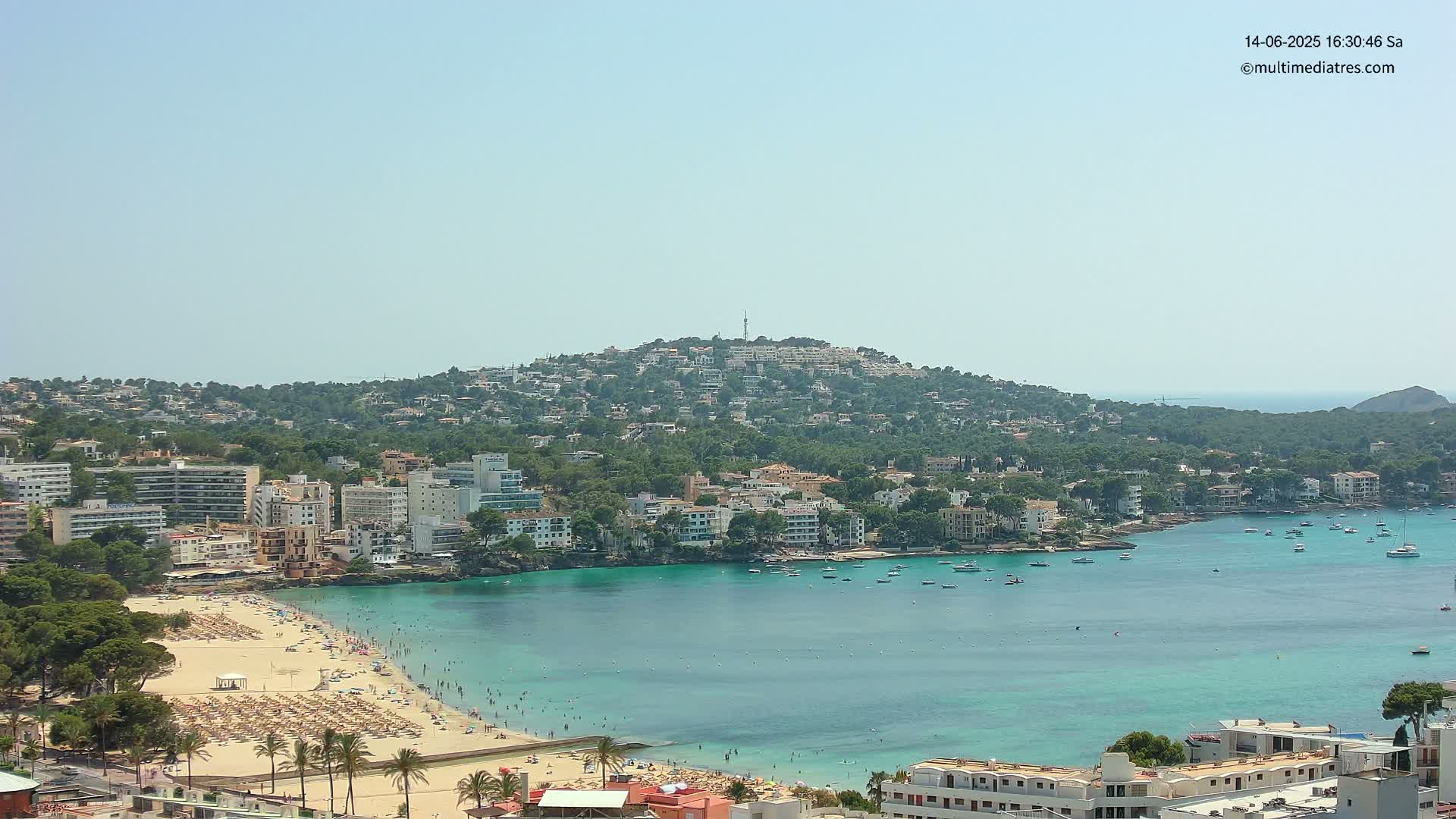 A coastal town nestled against a hillside, featuring a sandy beach with sunbathers, a calm turquoise bay with boats, and clear sunny skies.