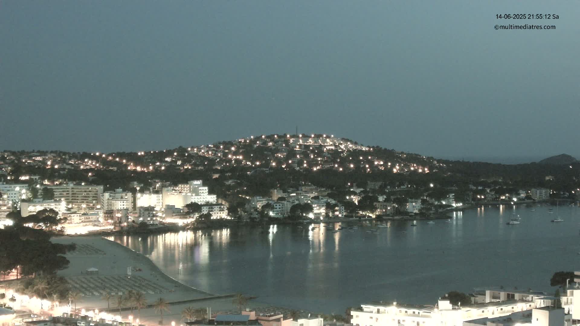 A coastal town nestled against a hillside, featuring a sandy beach with sunbathers, a calm turquoise bay with boats, and clear sunny skies.