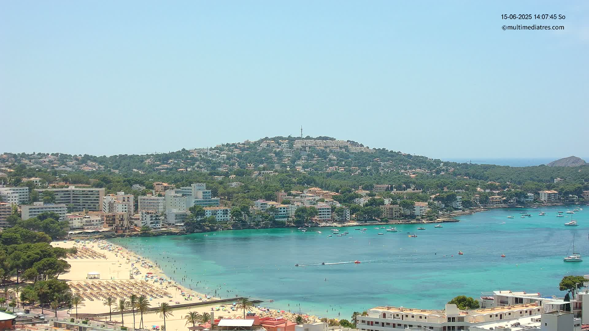 A sunny coastal town with a sandy beach crowded with people, numerous buildings lining the shore, and turquoise water filled with boats, all nestled against a green hill.