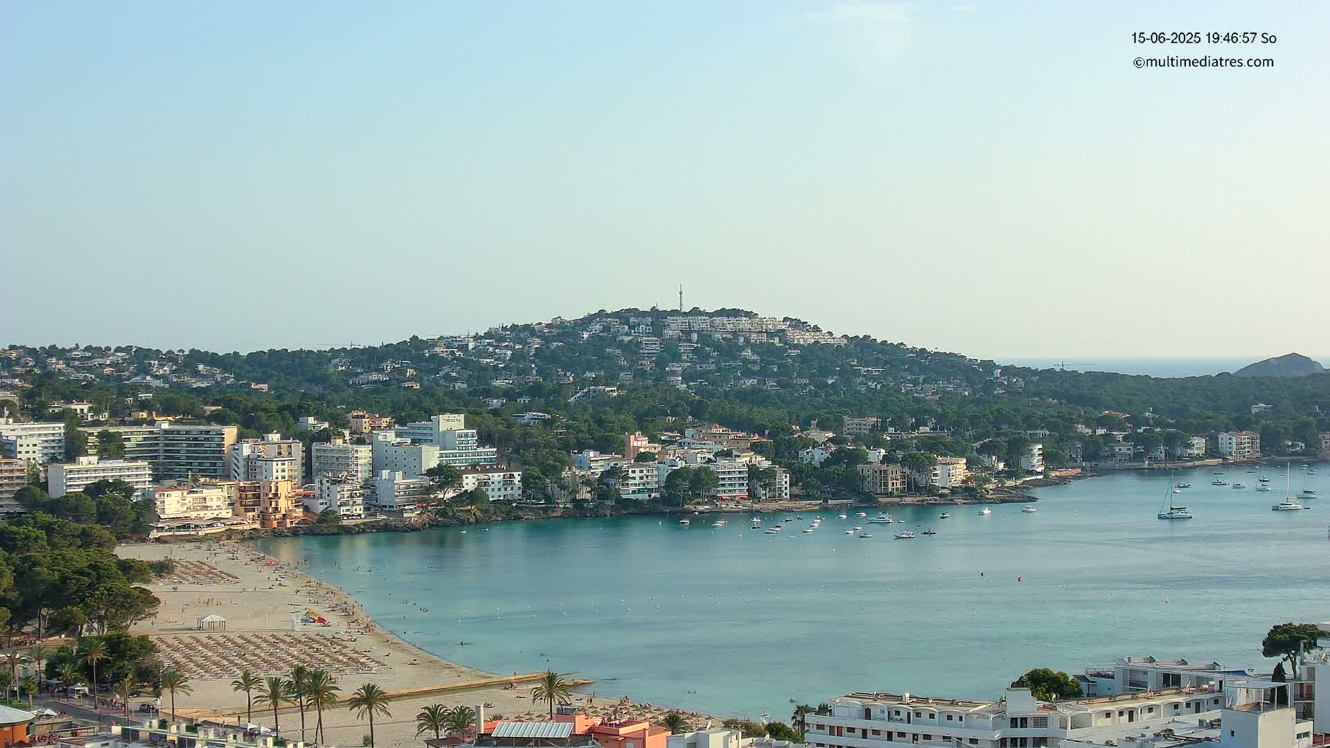 A coastal town with a sandy beach and turquoise bay nestled against a hill covered in buildings and trees under a clear sky.