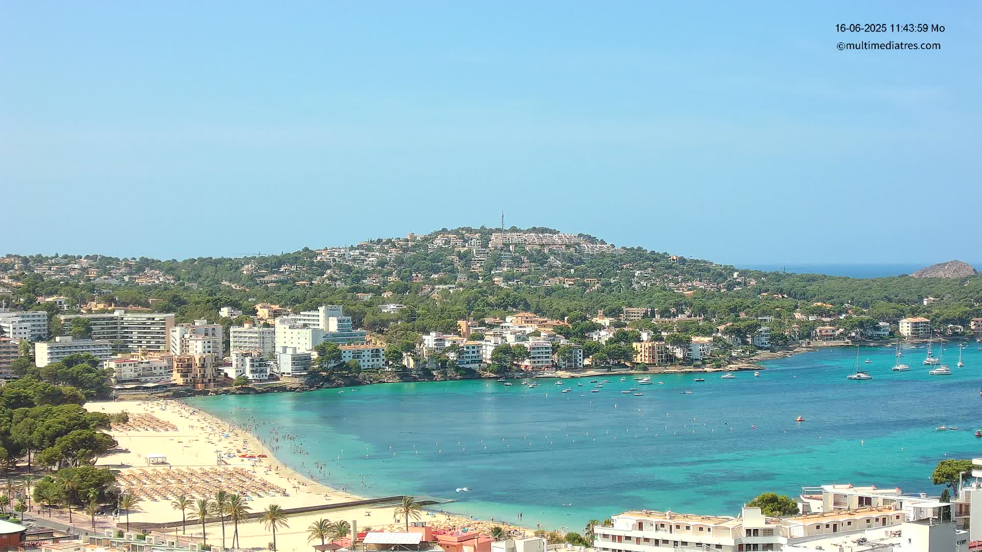 A sunny coastal town with a sandy beach, turquoise water filled with boats, and buildings covering a hillside.