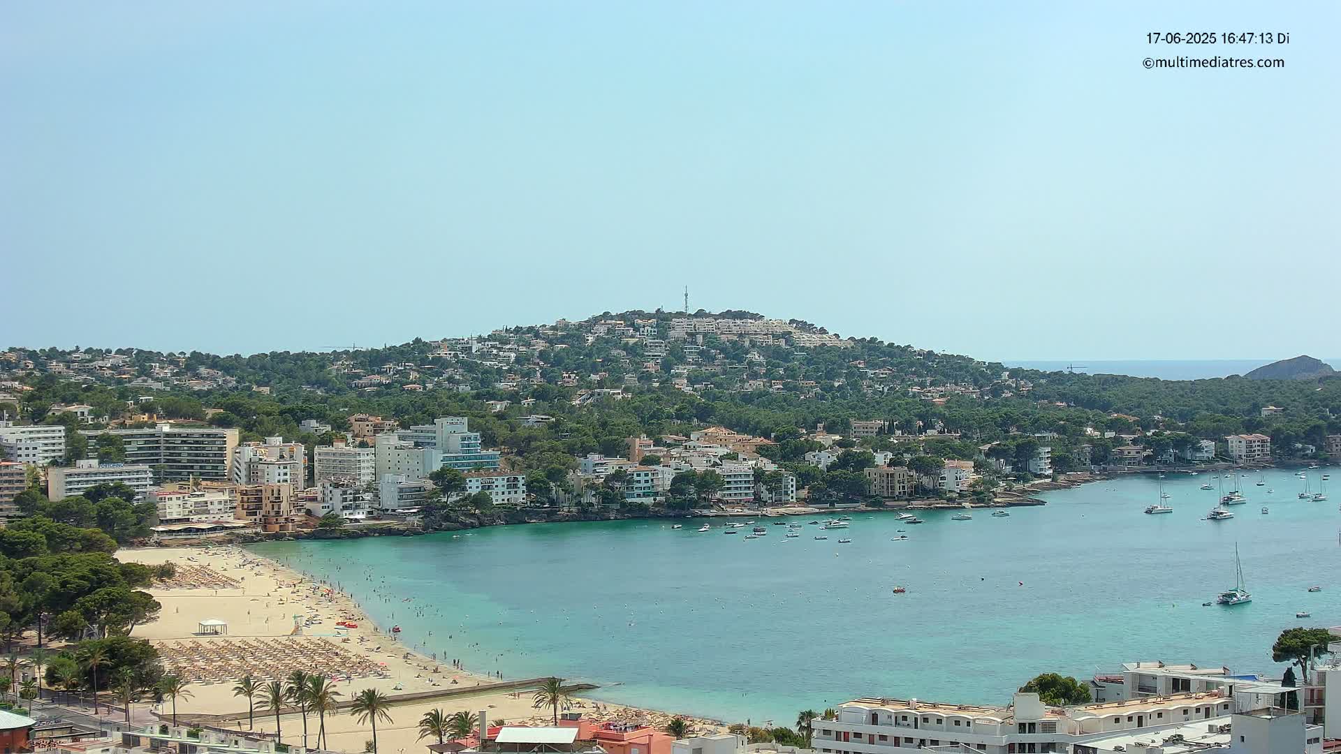 A sunny coastal town with a sandy beach and turquoise water, nestled below a hill covered in buildings and lush greenery.