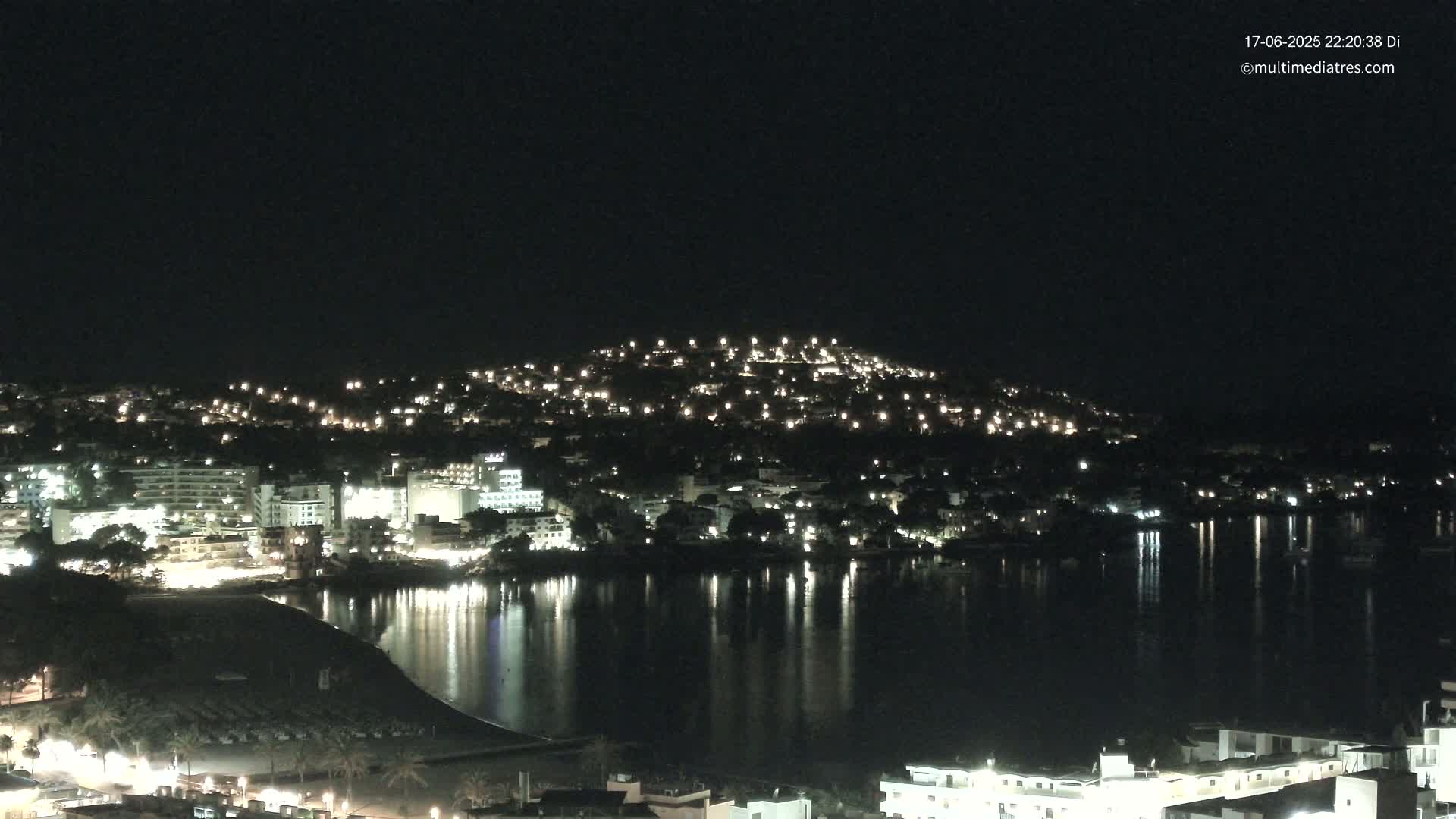 A nighttime view of a coastal town with many lights reflecting on a calm body of water.