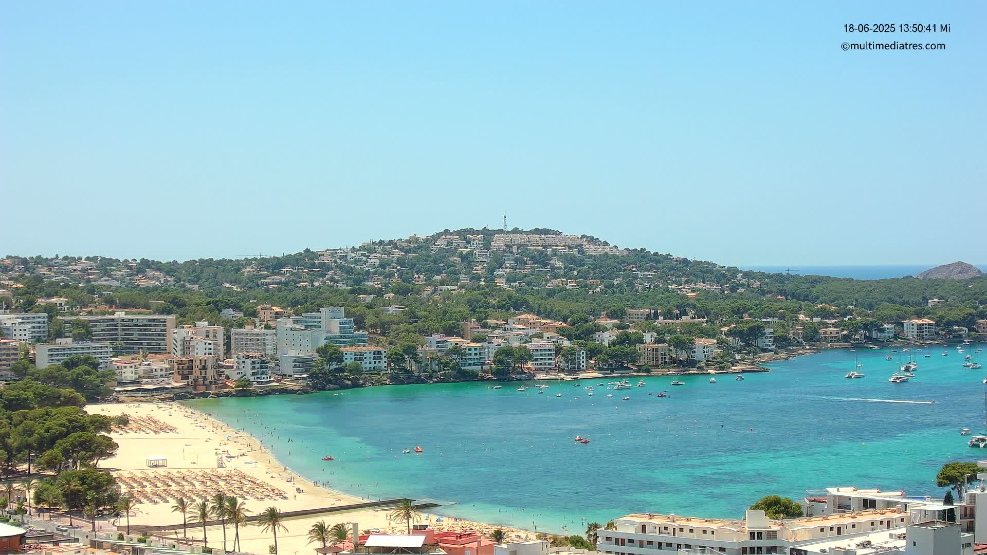 A sunny, clear day reveals a coastal town nestled on a hillside overlooking a turquoise bay with a sandy beach filled with people and umbrellas.