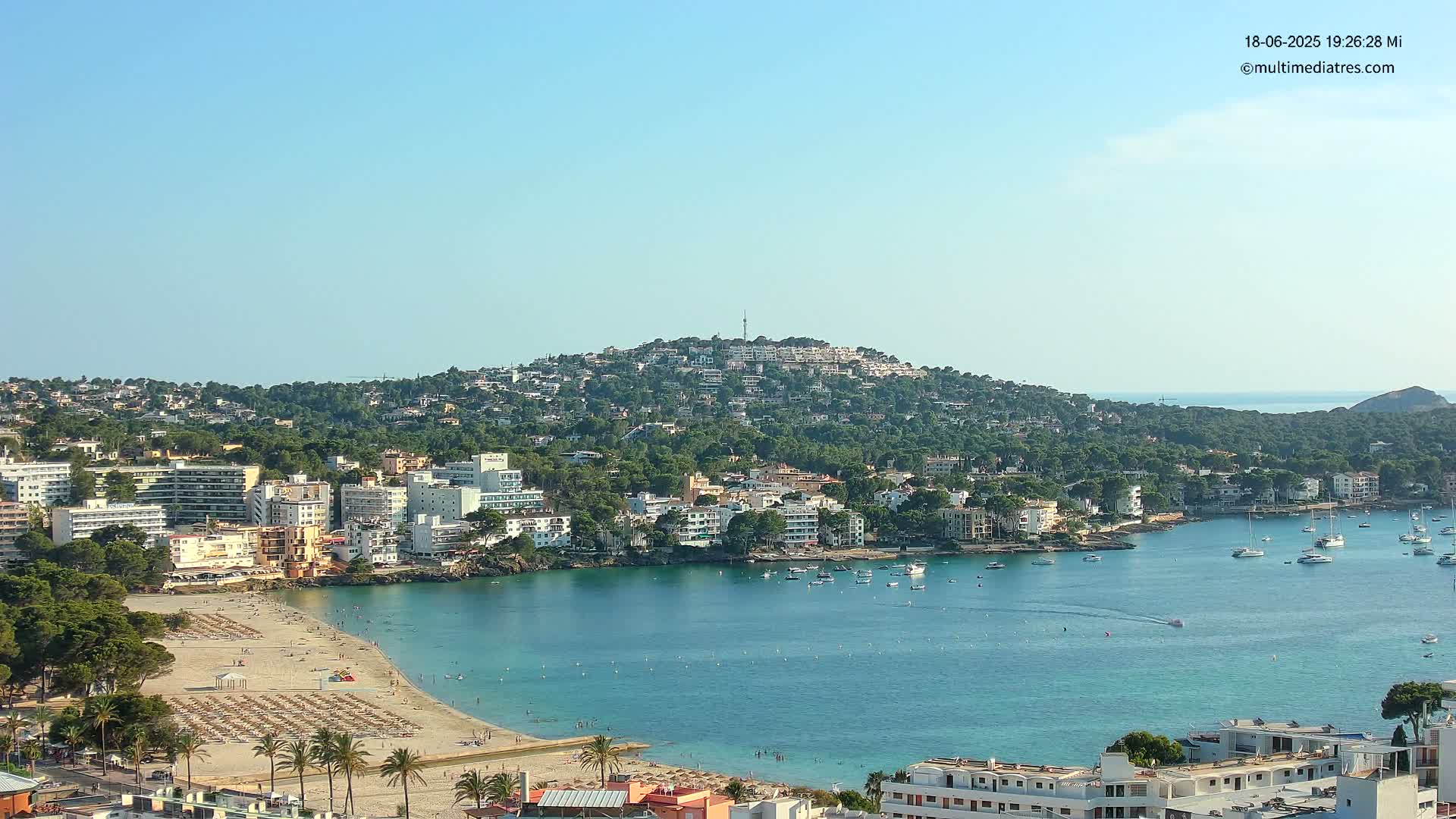 A coastal town with a sandy beach and turquoise water is nestled below a hill covered in houses and trees under a clear blue sky.