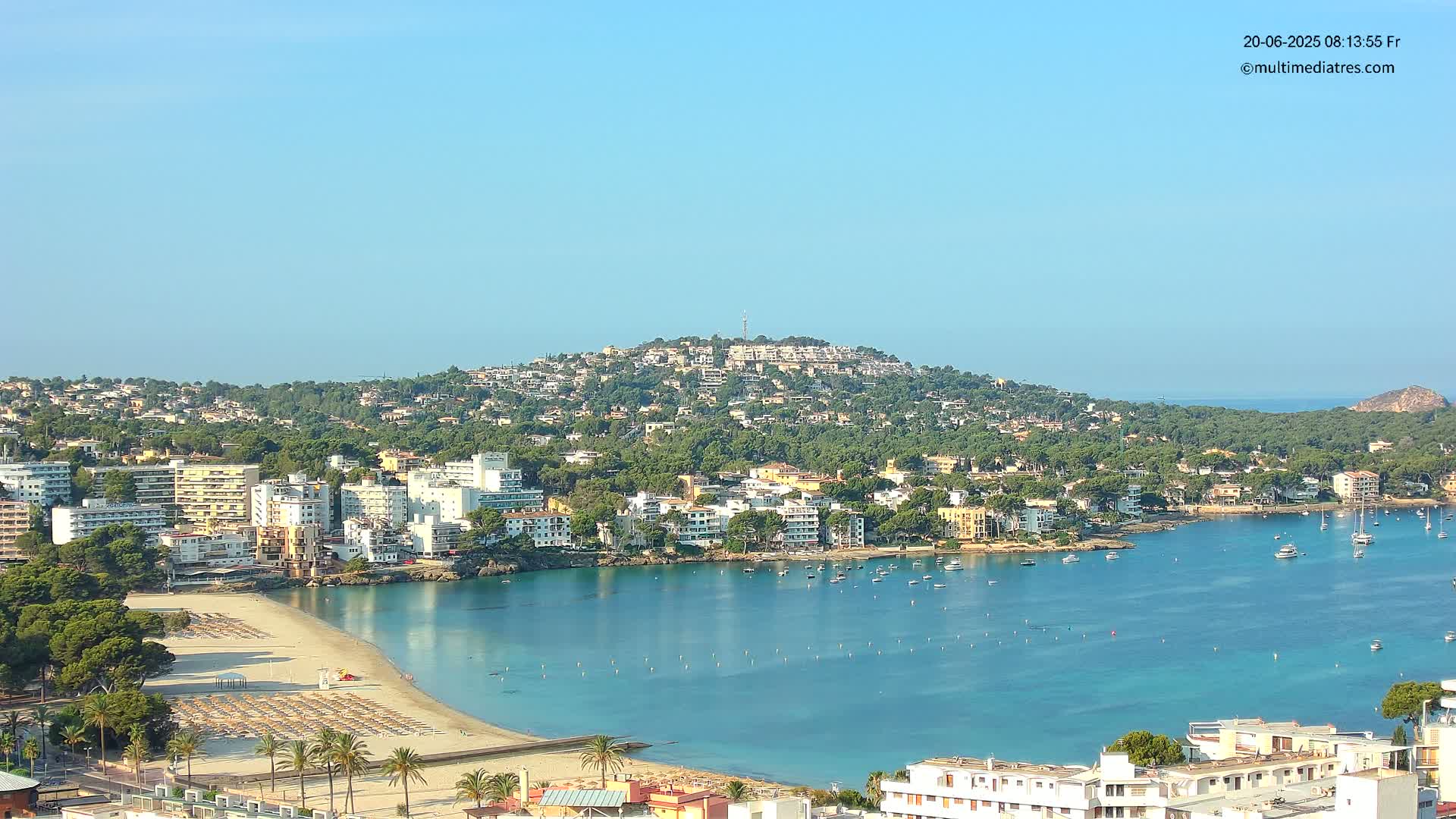 A coastal town with a sandy beach and calm, clear blue water is nestled beneath a hill covered in buildings and lush green vegetation under a bright, clear sky.