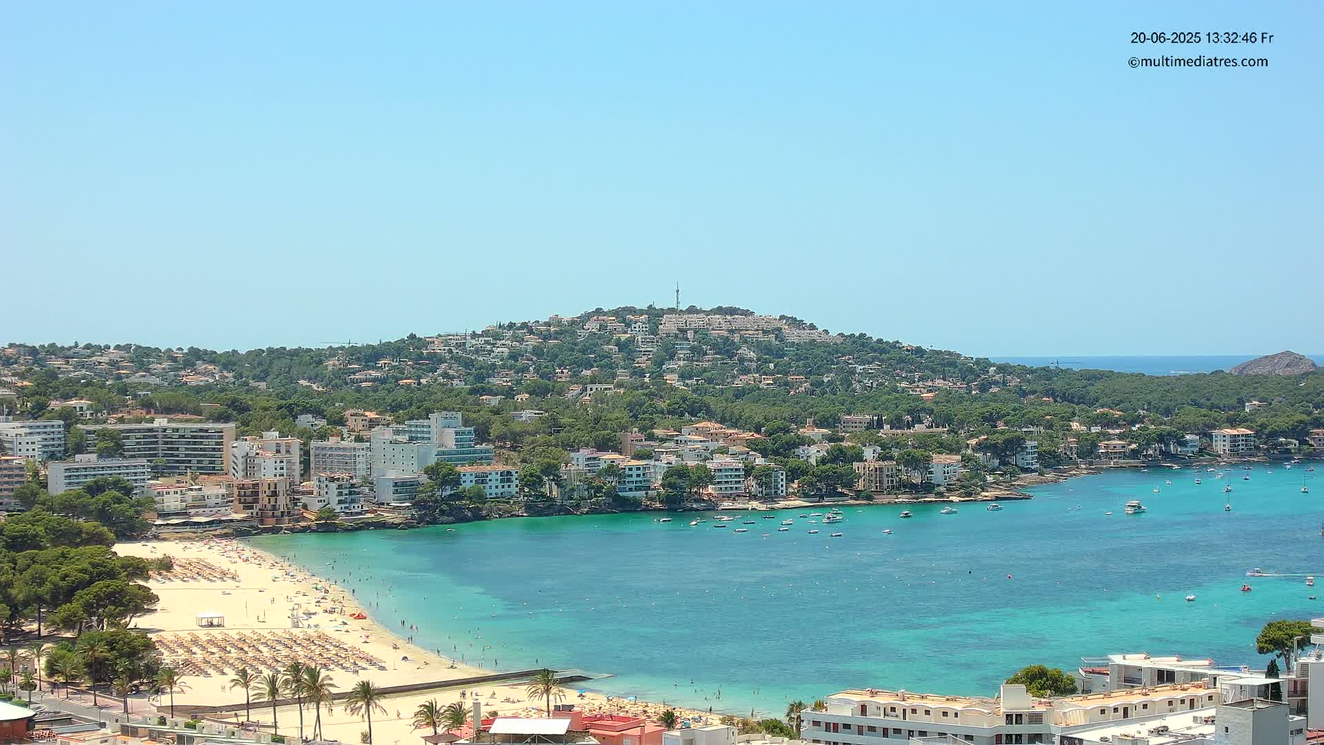 A clear, sunny day reveals a coastal town nestled around a turquoise bay, with a hill covered in buildings and greenery in the background.
